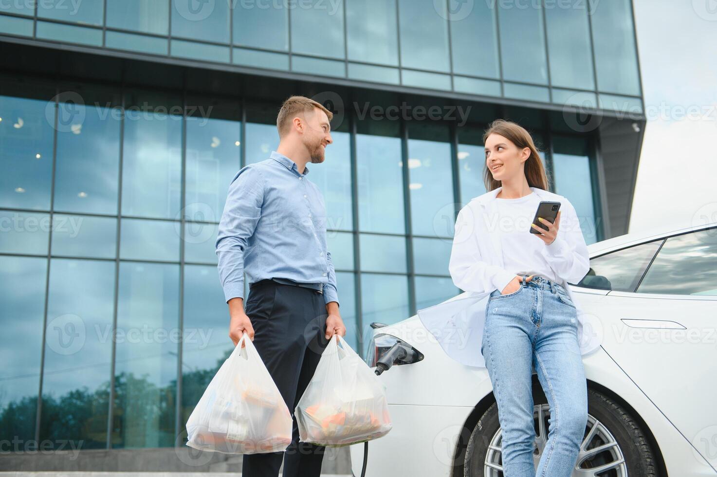 Smiling man and woman on the charging station for electric cars. A man is charging a car, a woman is standing with a phone photo