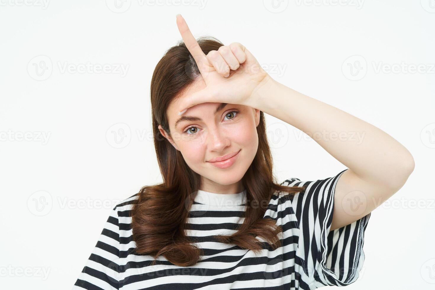 Close up portrait of young brunette girl mocking you, showing loser finger gesture, l letter with hand on forehead, smiling with pleased face, standing isolated on white background photo