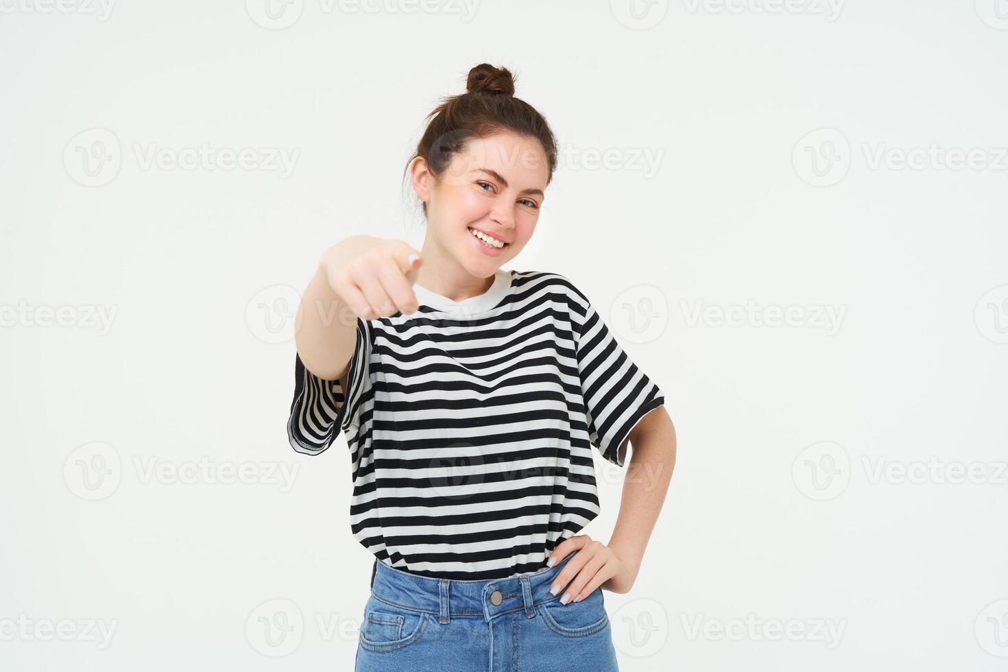 Image of confident, happy young woman in casual clothes, pointing finger at camera, laughing and smiling, standing over white background photo