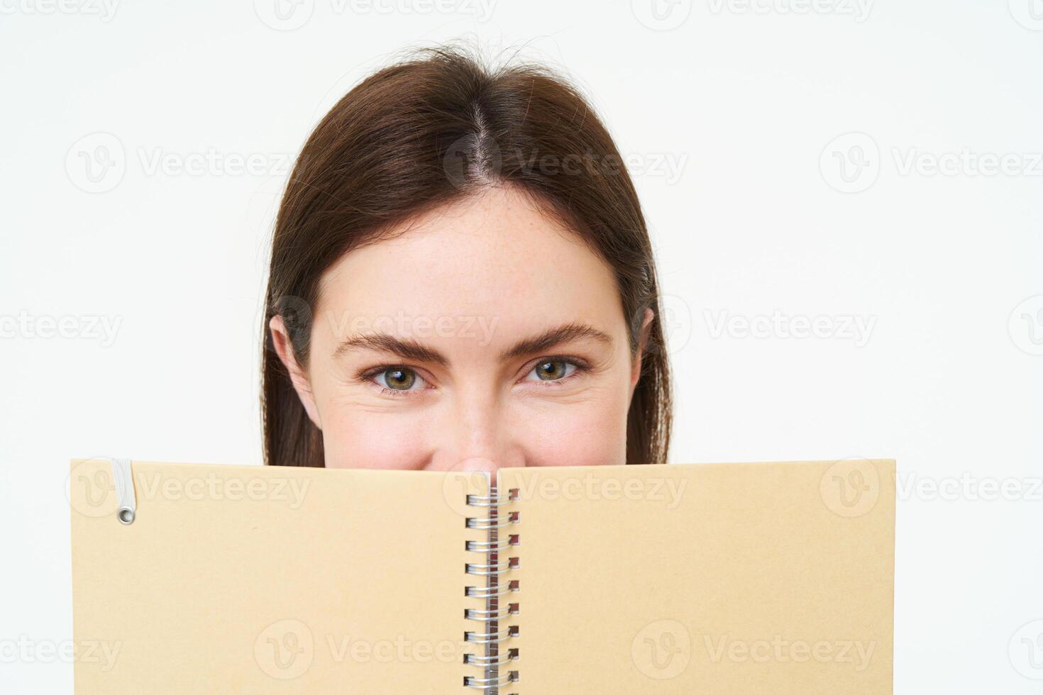 Portrait of cute young woman hides her face behind planner, holds notebook against her face and smiles, isolated over white background photo
