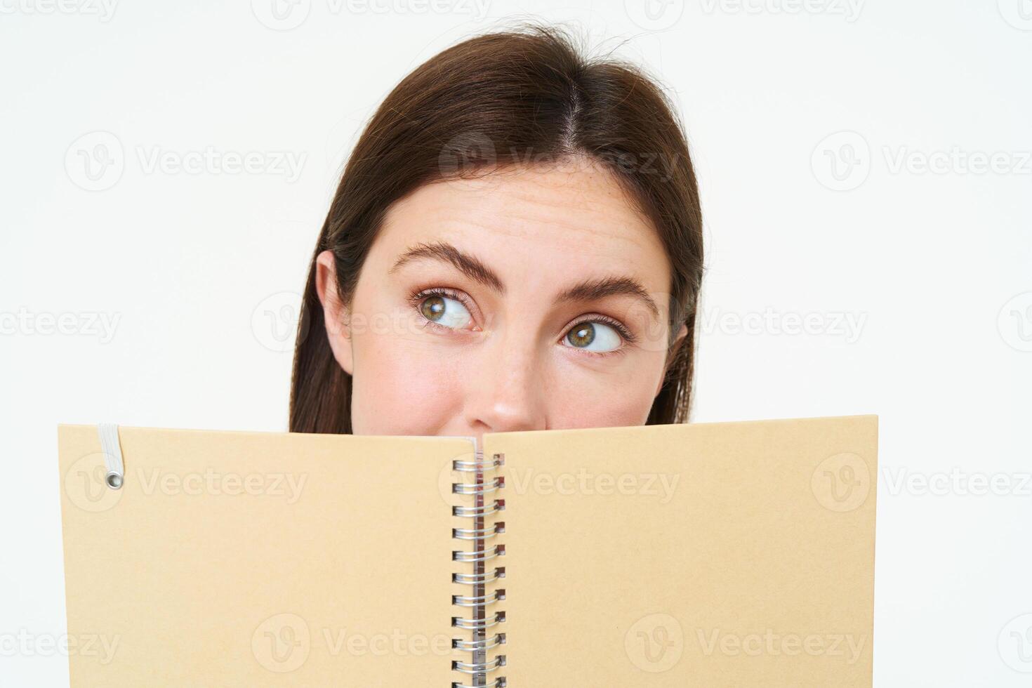 joven mujer sostiene cuaderno diario planificador siguiente a su rostro, escritura abajo tarea, haciendo notas, mirando considerado, en pie terminado blanco antecedentes foto