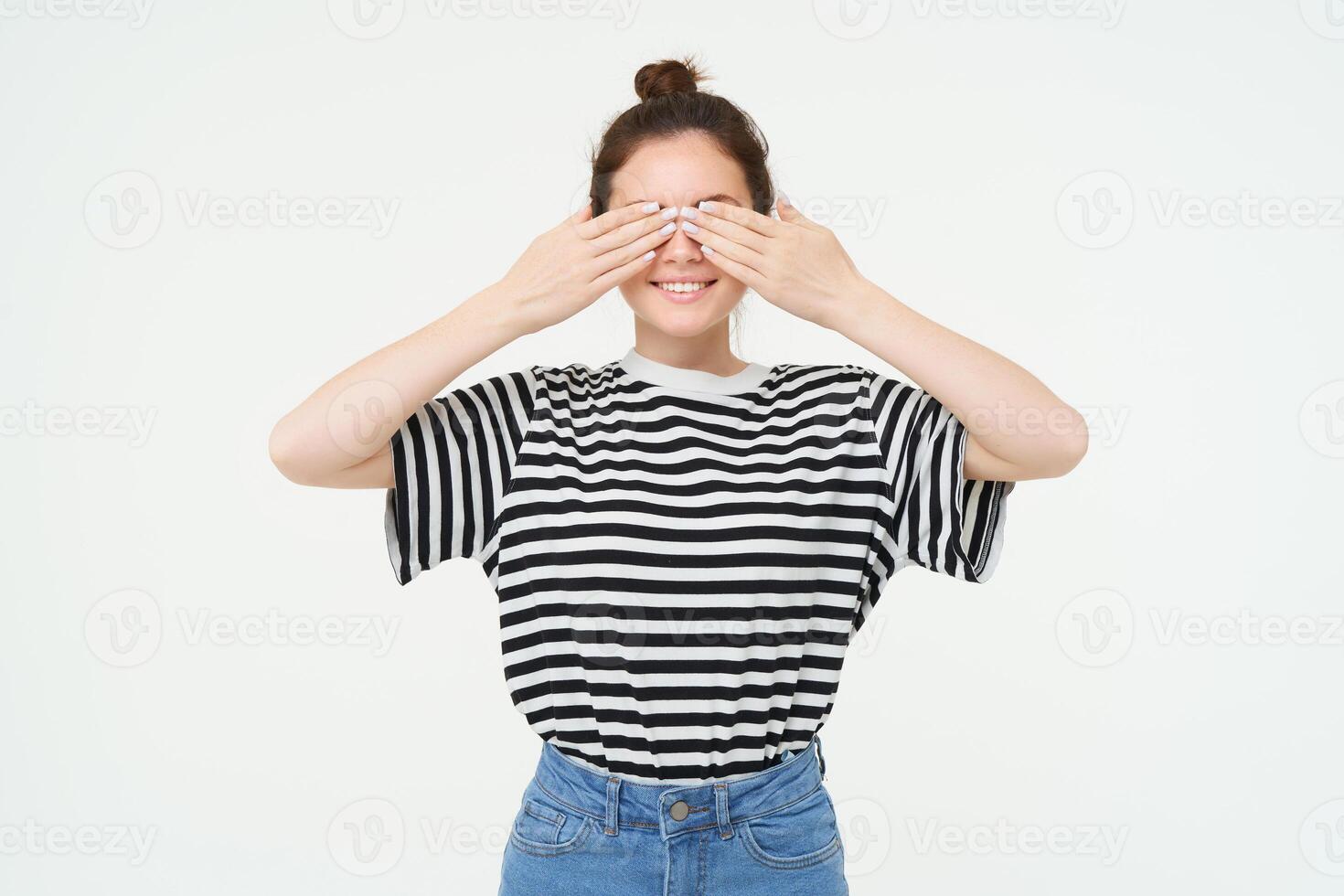 Portrait of girl waiting for surprise, covers eyes with hands, peeking through fingers, stands over white background photo