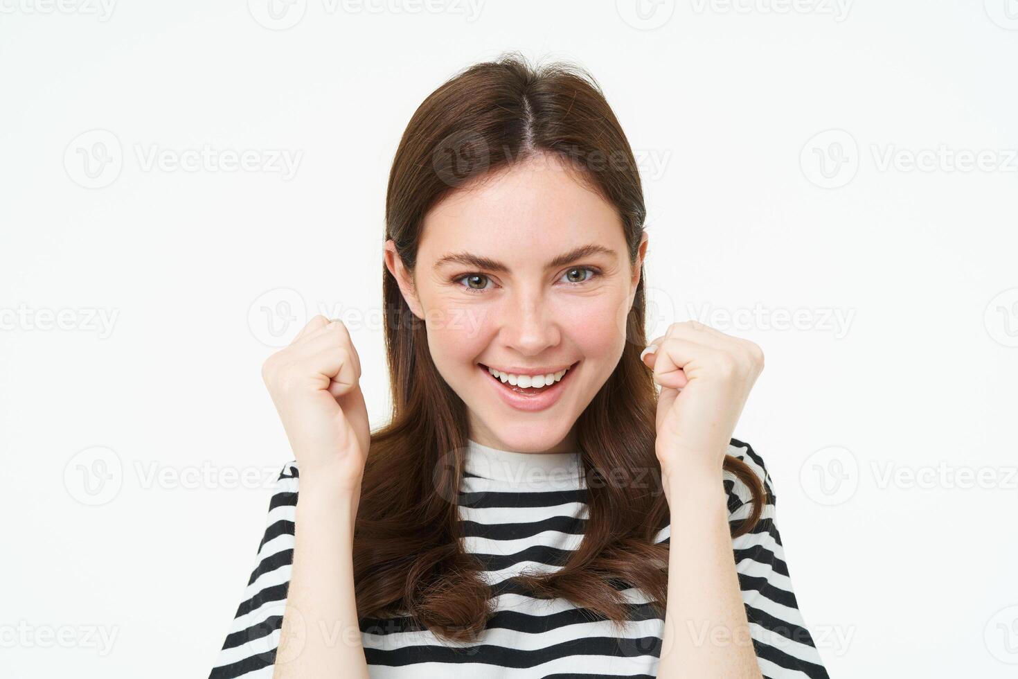 Image of young woman looking with pleased, satisfied face at camera, winning, shaking hands in triumph, celebrating victory, achievement, standing over white background photo