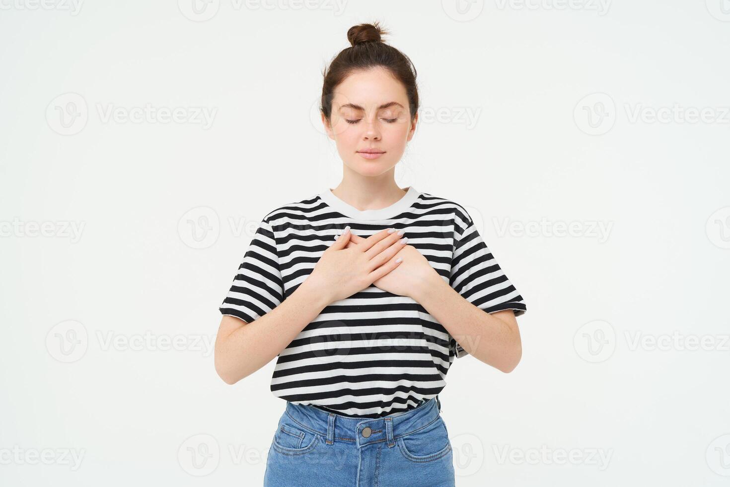 Portrait of young woman with calm, relaxed face, holds hands on heart with eyes closed, stands over white background photo