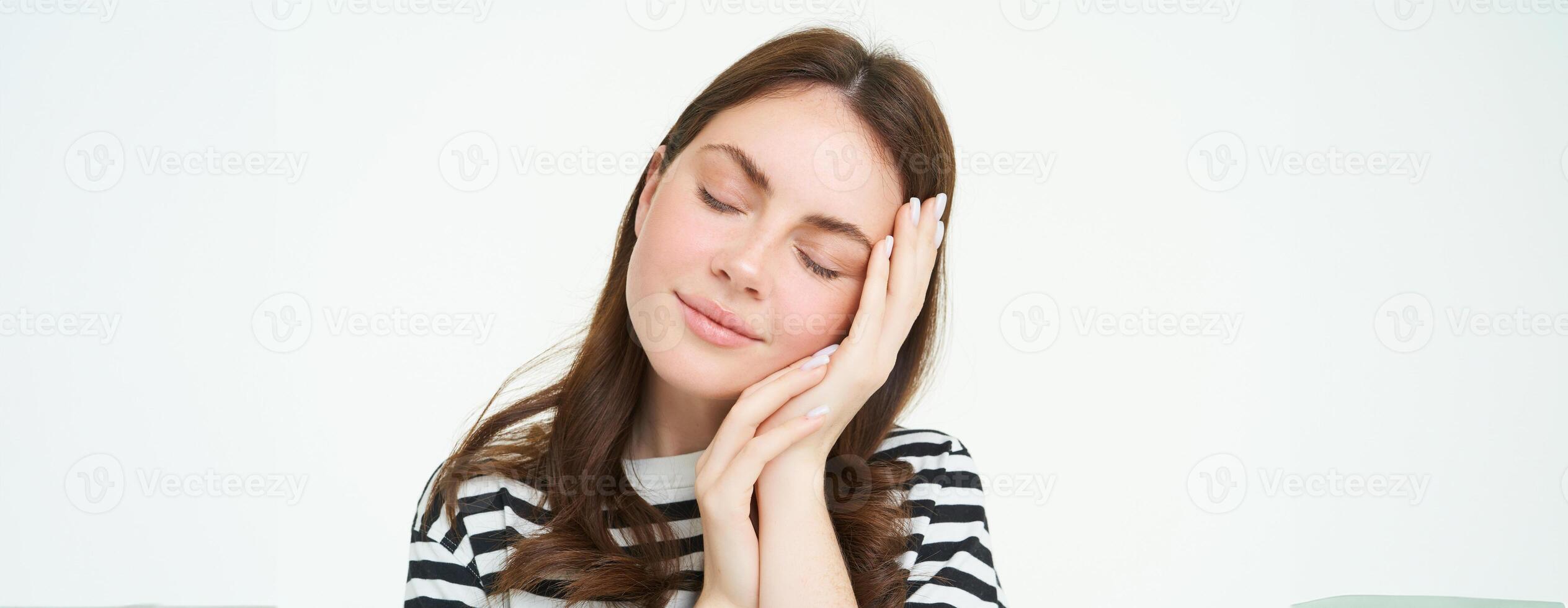 Close up portrait of tender, smiling young woman, closes her eyes and touches her palm, sleeping on her hand, isolated on white background photo