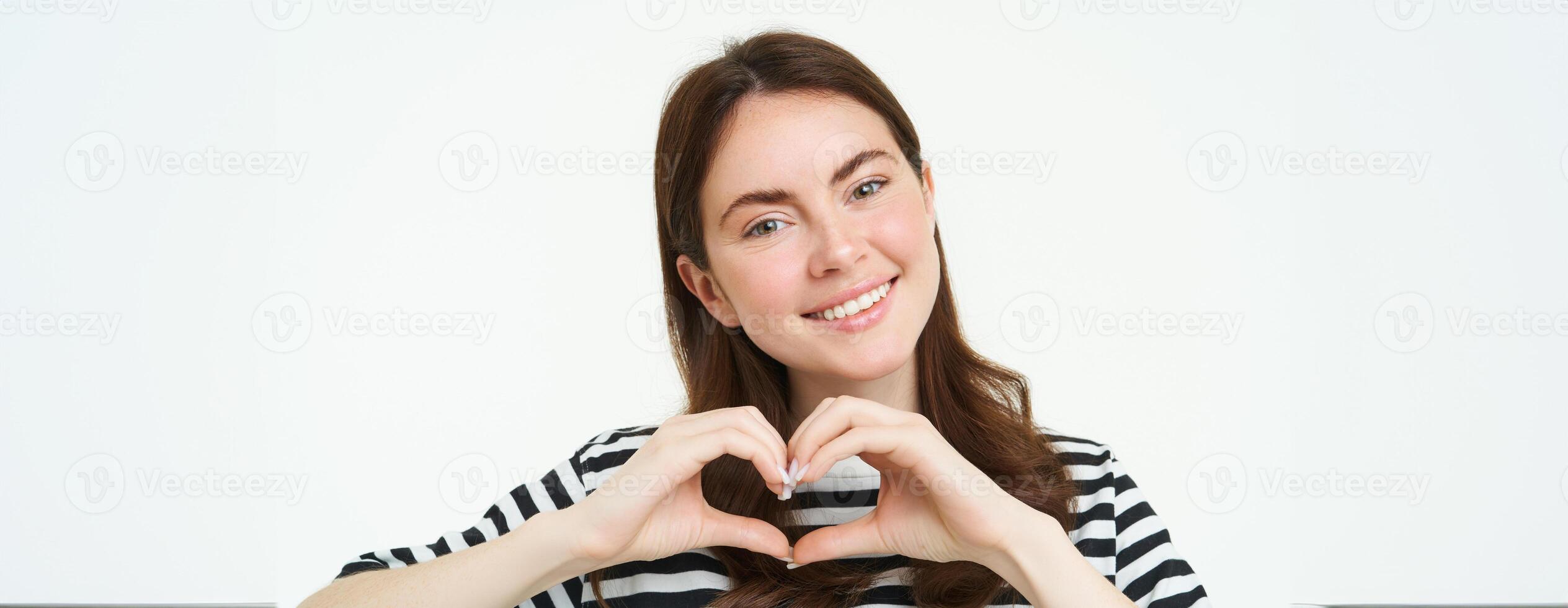 Portrait of smiling, happy girlfriend shows heart with hands and looking with care at camera, express her love or affection, like product, standing over white studio background photo
