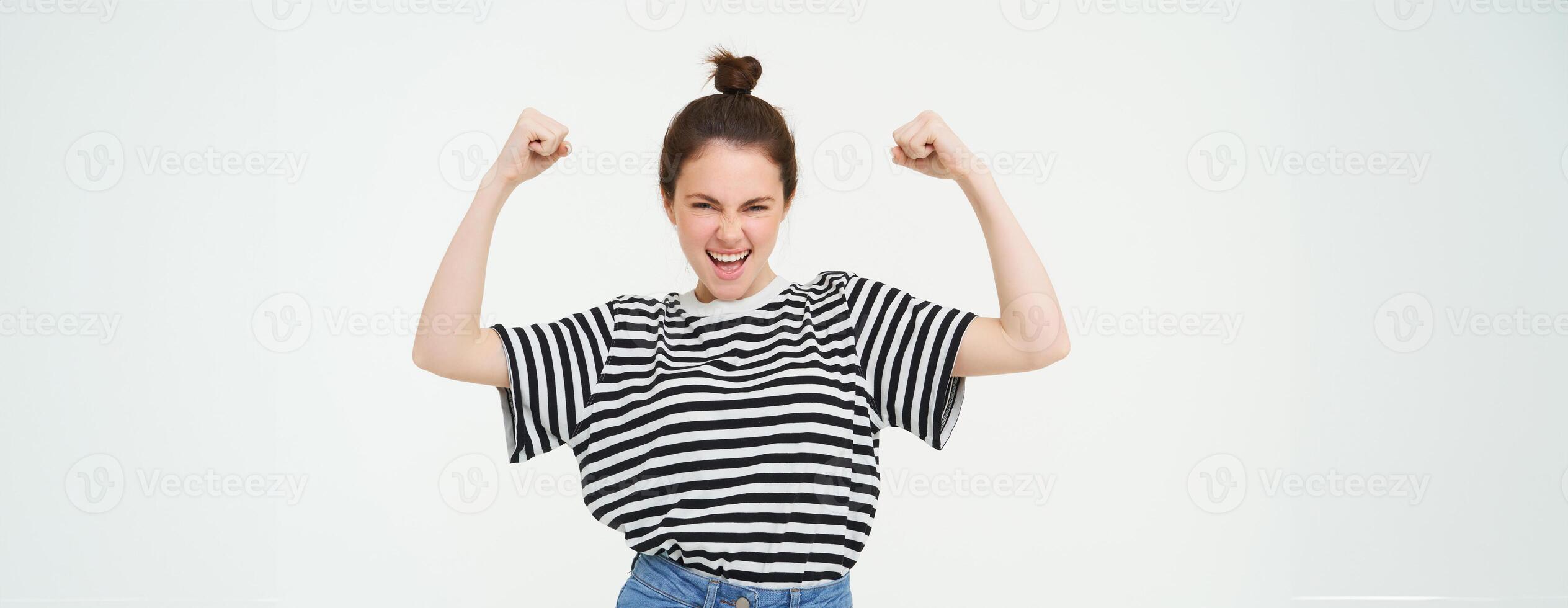 Image of sassy, strong and confident woman shows her muscles, flexing biceps, raising her arms high, standing over white background photo