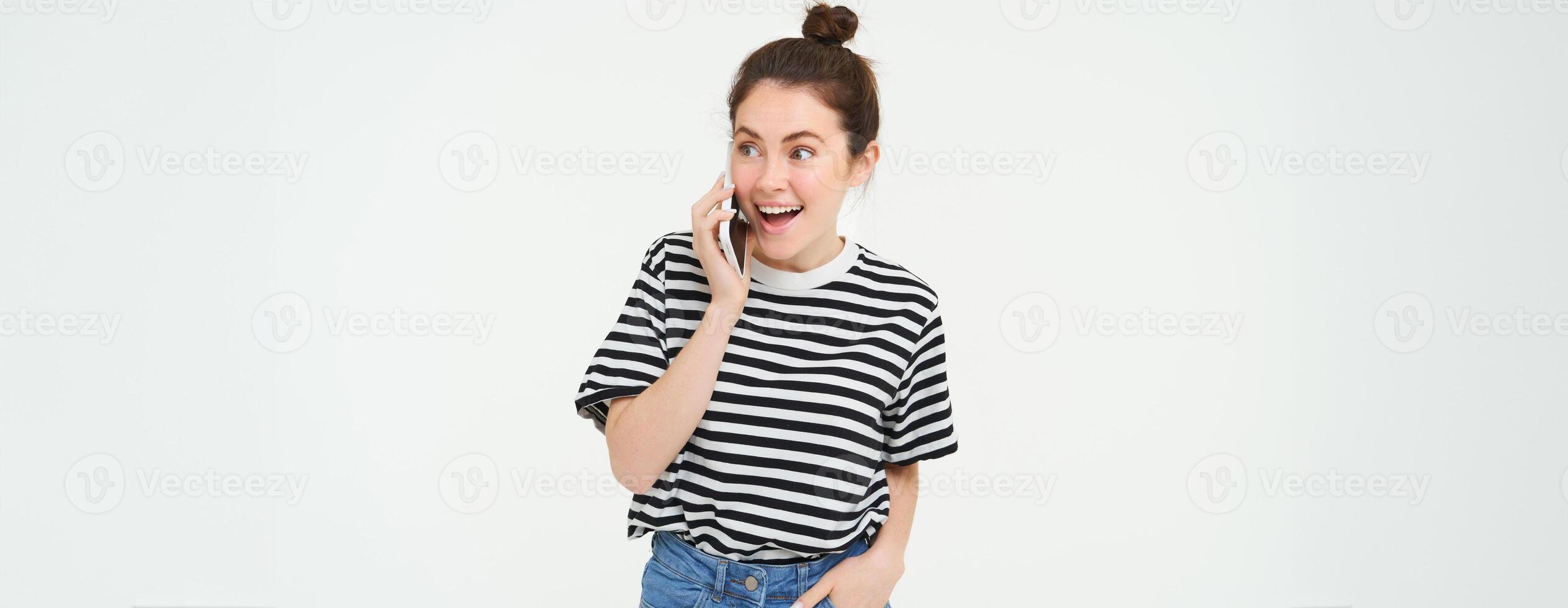 Portrait of enthusiastic young woman talking on mobile phone, chatting with friend, reacts to surprising news received by telephone, white background photo