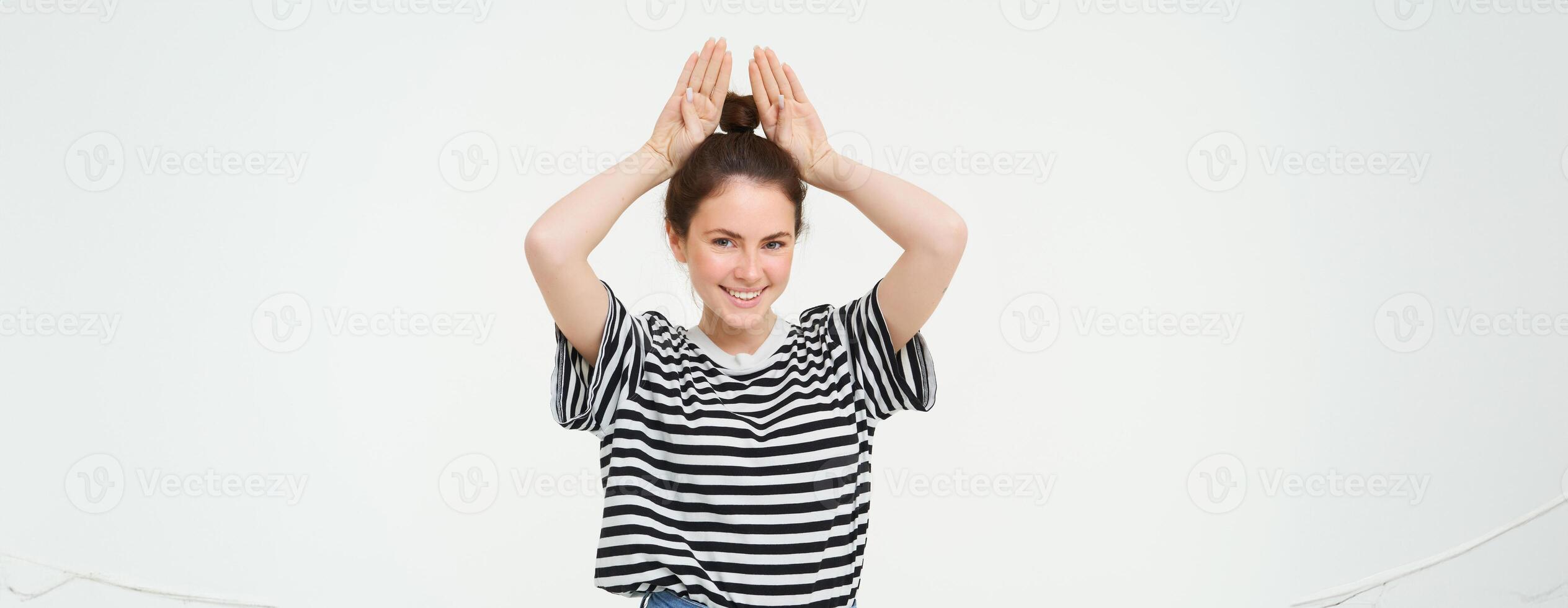 Image of beautiful, happy young woman showing bunny ears on top of her head, looking excited, posing over white background photo