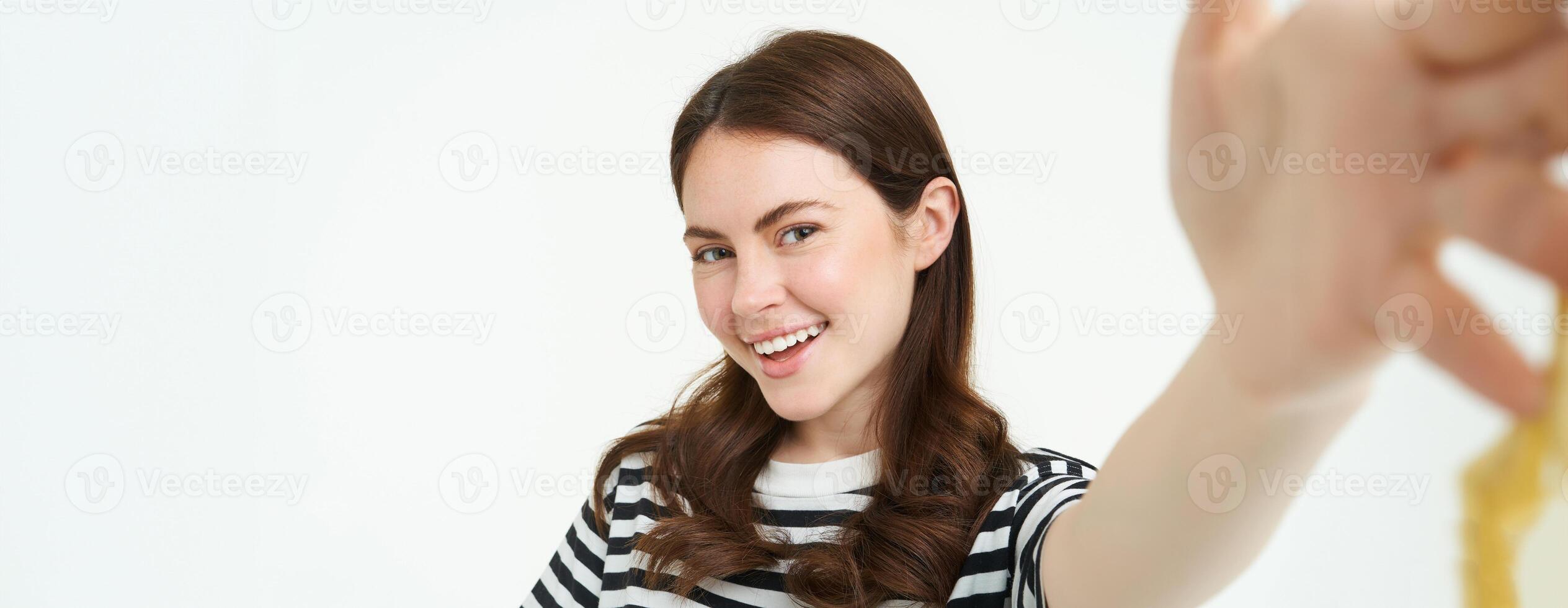 Portrait of beautiful young woman, 25 years old, holds smartphone, takes selfie, posing for a photo, standing over white background photo