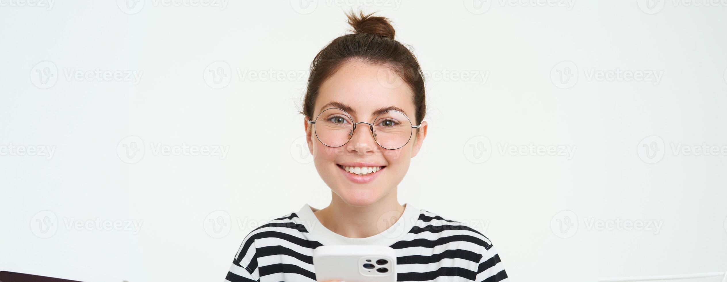 Portrait of beautiful young woman in eyewear, holding smartphone, using mobile phone, online shopping in application, standing over white background photo