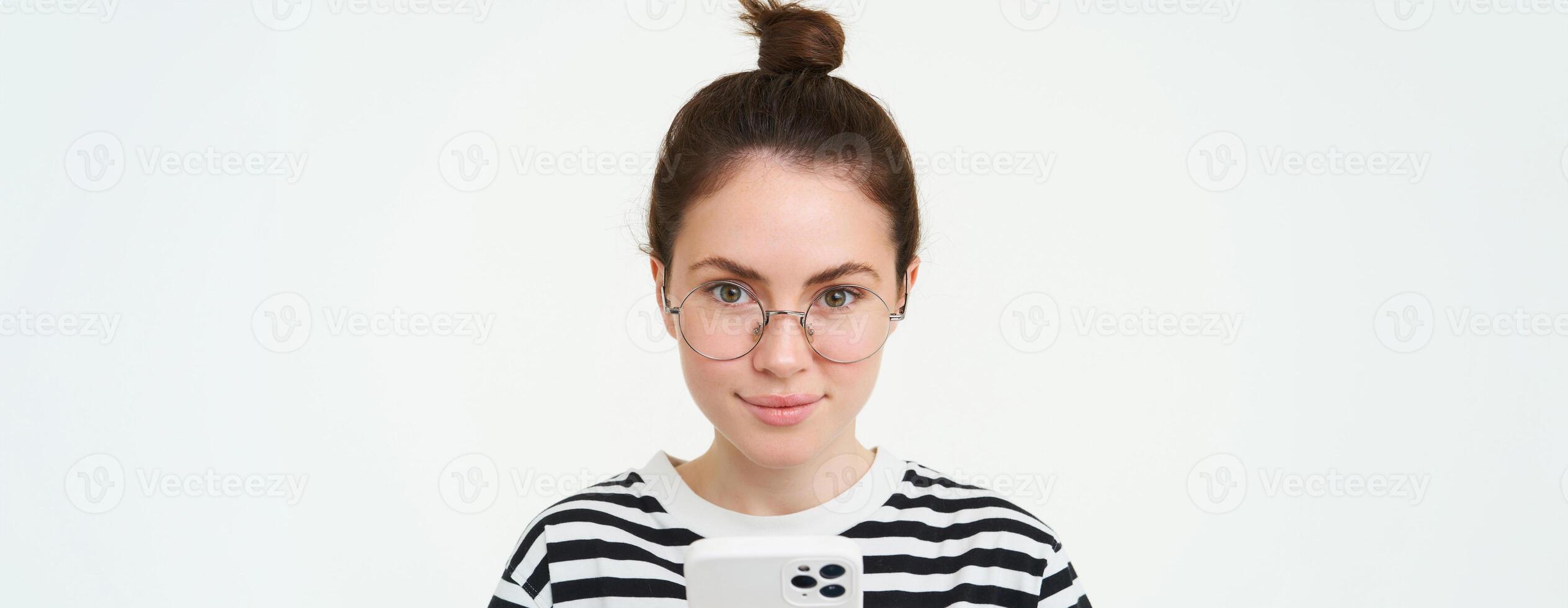 Portrait of beautiful young woman in eyewear, holding smartphone, using mobile phone, online shopping in application, standing over white background photo