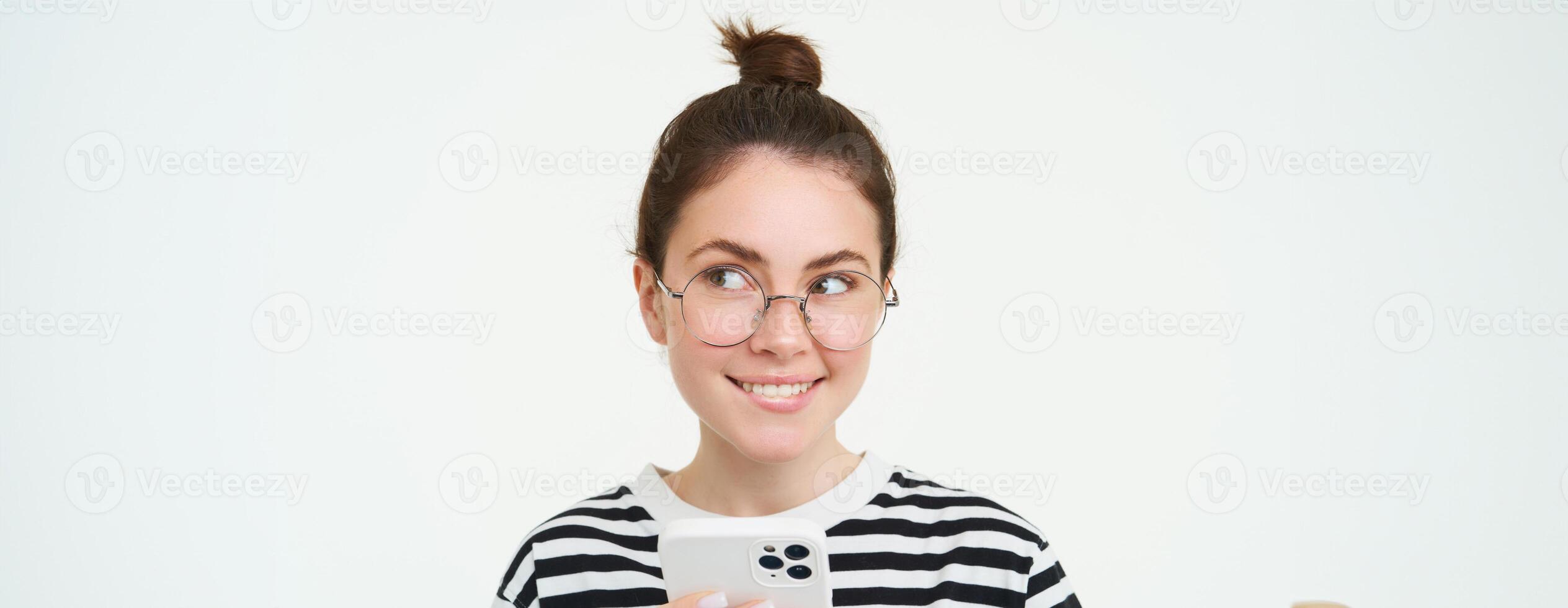 Image of young woman in glasses, using her mobile phone, standing with smartphone and smiling, standing over white background photo