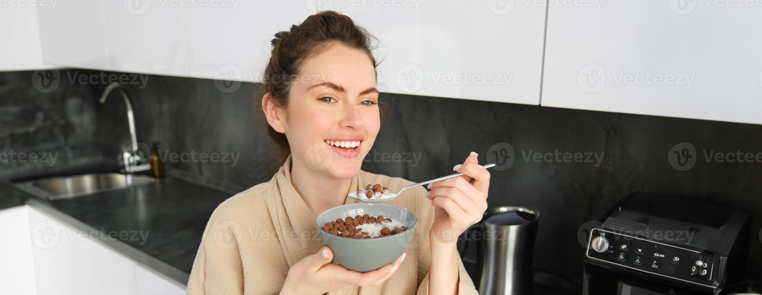 Good-looking brunette woman eating her breakfast, standing in kitchen near worktop and holding bowl of cereals with milk, enjoying her morning, wearing cosy bathrobe photo