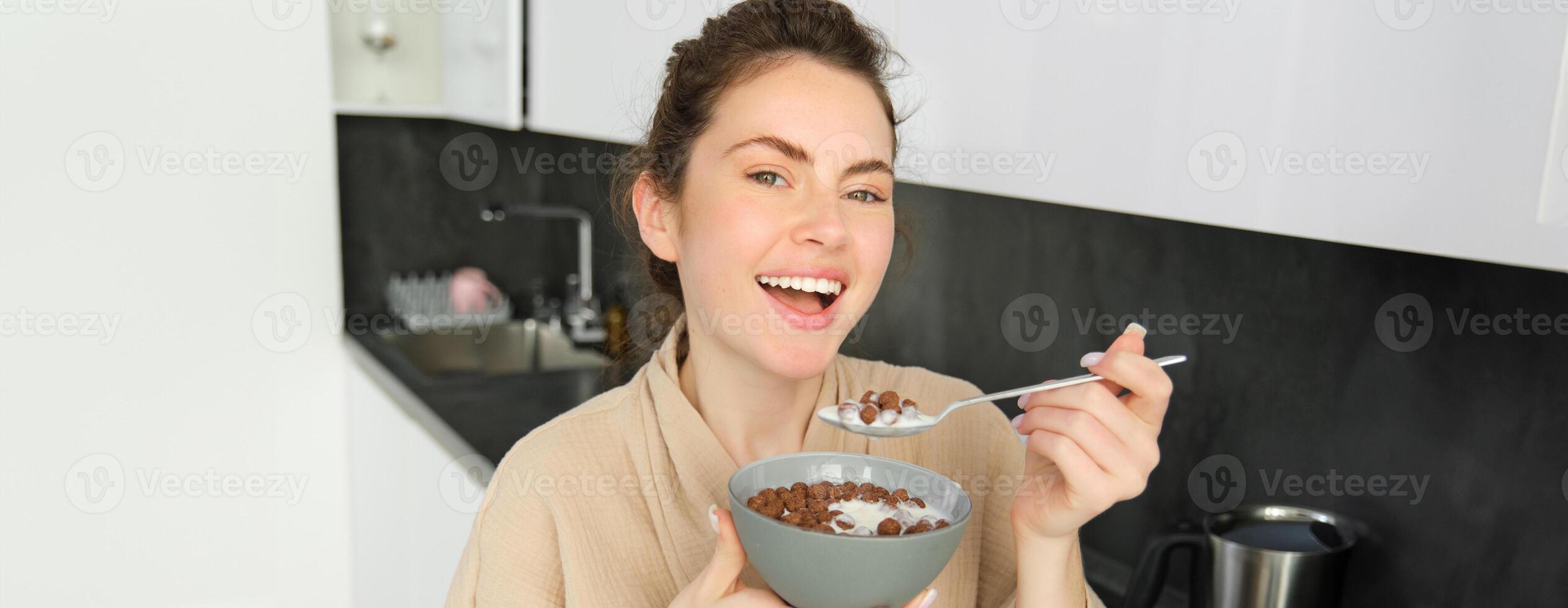 Happy mornings. Gorgeous young woman eating cereals with milk, standing in kitchen with breakfast bowl, enjoying start of the day, smiling photo