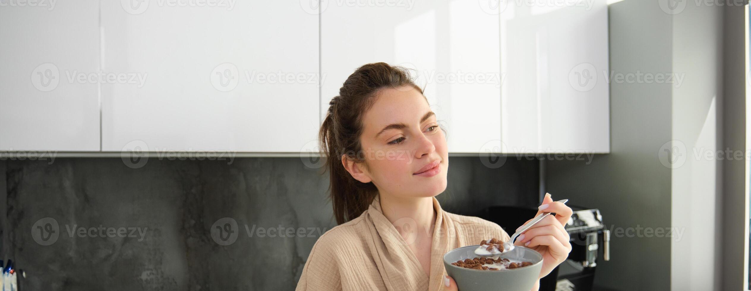 Happy mornings. Gorgeous young woman eating cereals with milk, standing in kitchen with breakfast bowl, enjoying start of the day, smiling photo