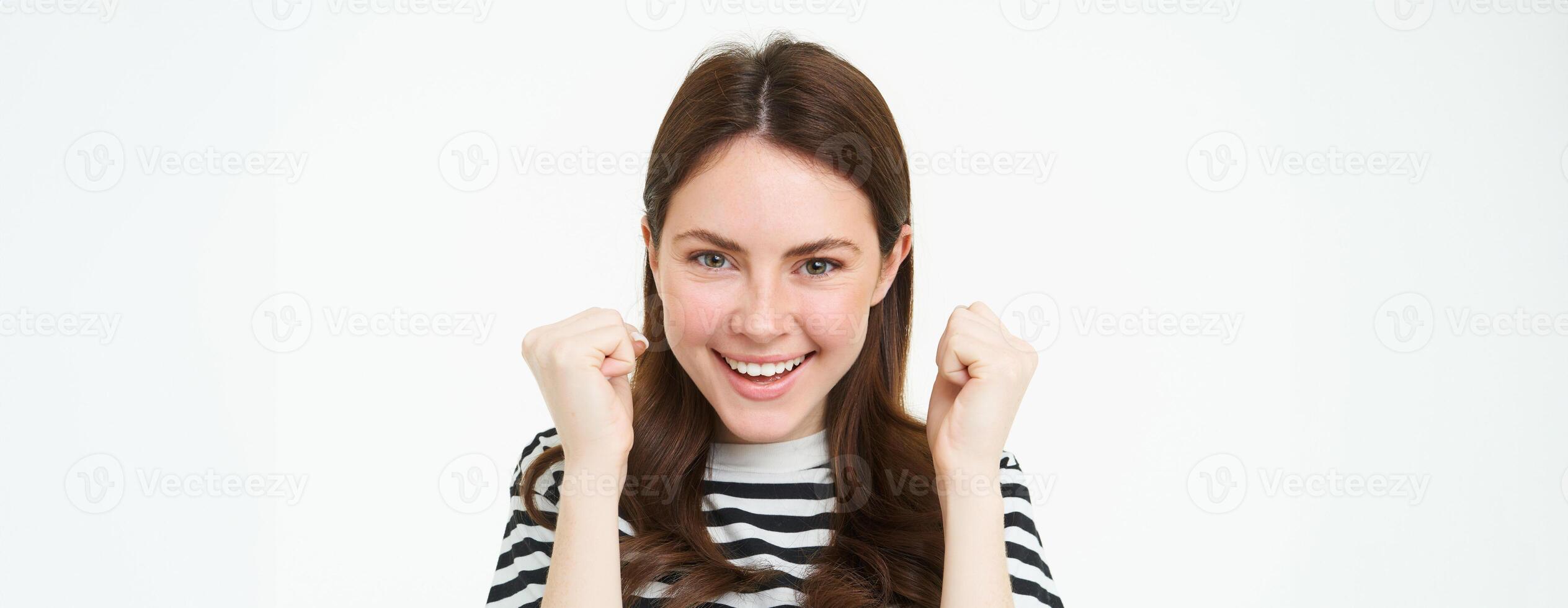 Image of young woman looking with pleased, satisfied face at camera, winning, shaking hands in triumph, celebrating victory, achievement, standing over white background photo