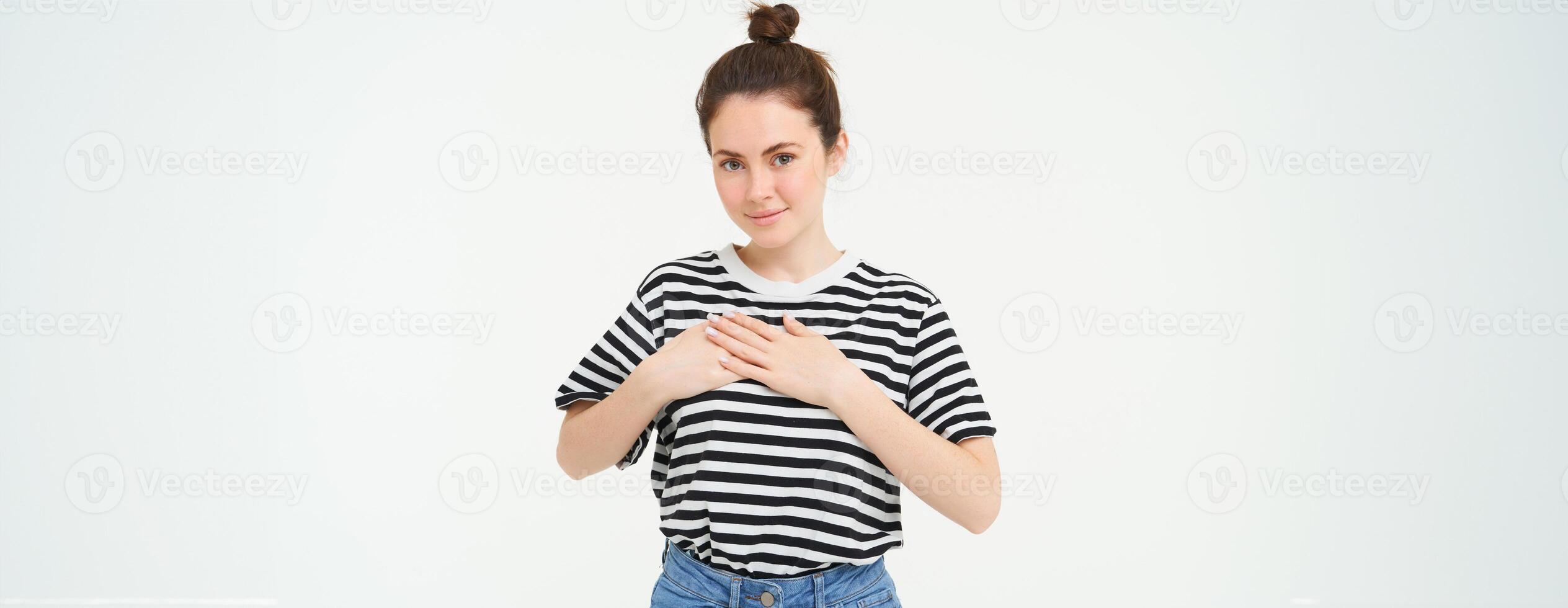 Young smiling woman, feels gratitude, thankful for something, holds hands on heart and looks tender at camera, standing over white background photo