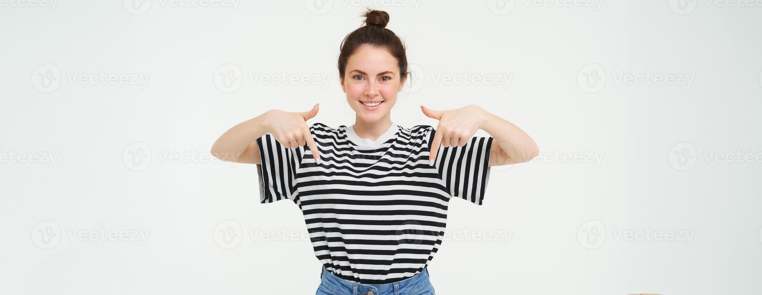 Portrait of beautiful, smiling young female model, showing advertisement, pointing fingers down, standing over white background photo