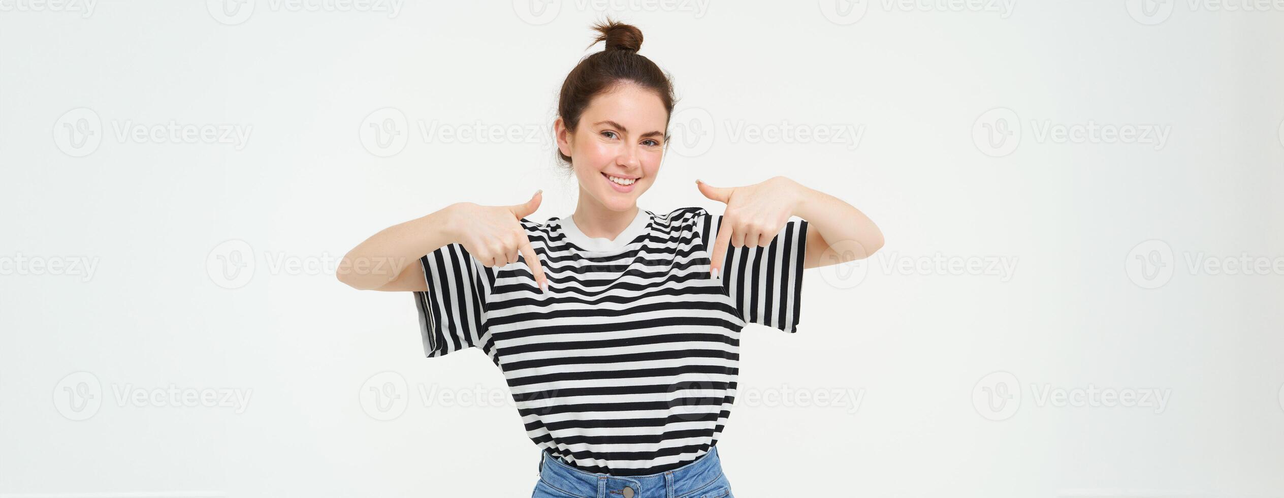 Portrait of smiling, beautiful young woman, pointing fingers down, showing advertisement, posing against white background photo