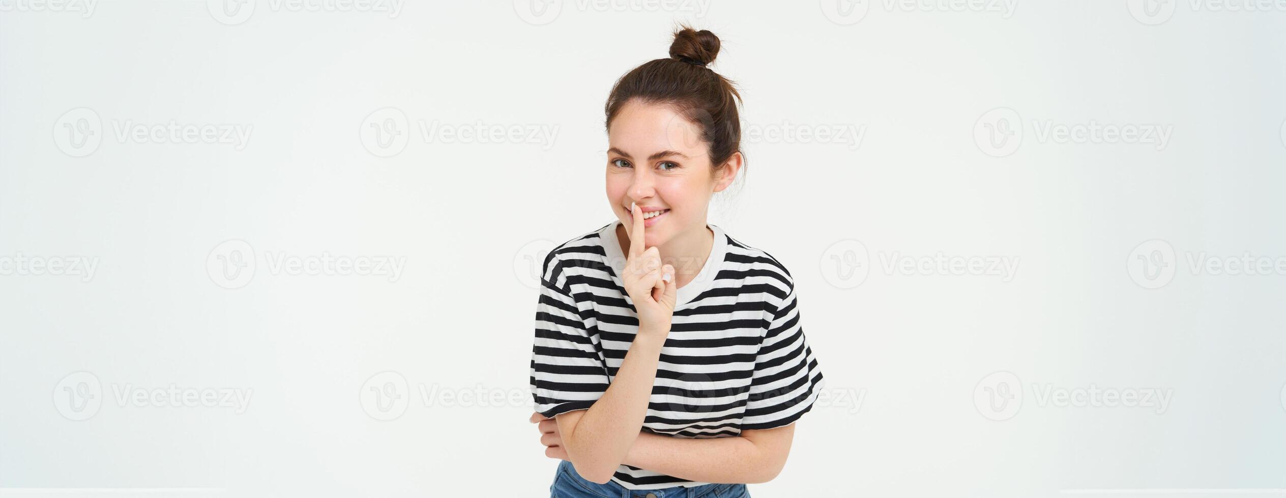 Beautiful, coquettish girl, holds finger on lips, promise to keep secret, shh gesture, hush sign, stands over white background photo