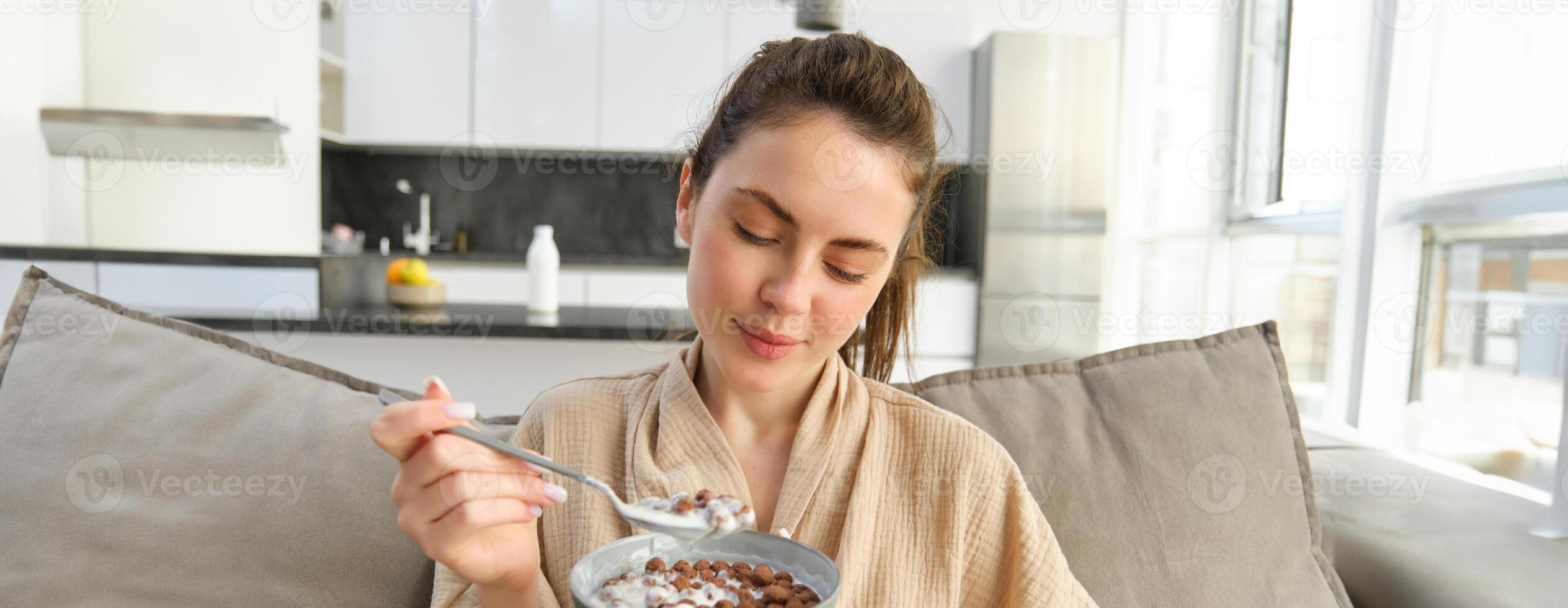 Image of smiling, happy young woman eating breakfast, holding bowl of cereals with milk, having meal at home photo