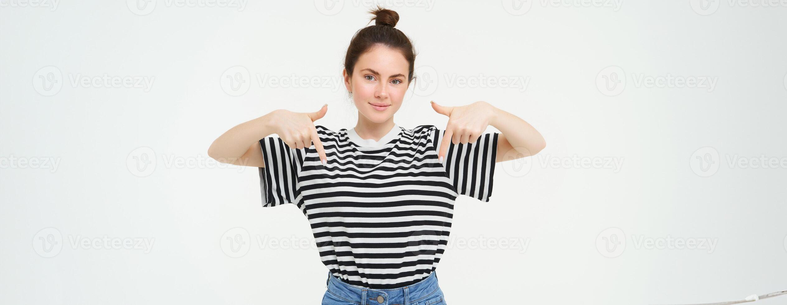 Portrait of confident, smiling brunette woman, pointing fingers down, showing banner on bottom, follow link below gesture, standing over white background photo