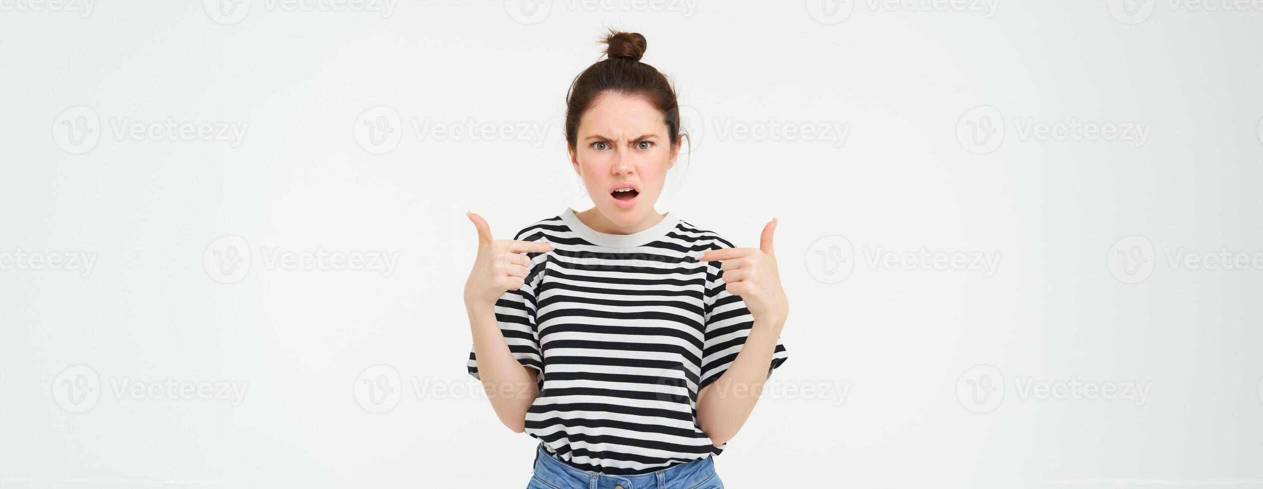 Annoyed woman pointing at herself and frowning, arguing, looking frustrated and disappointed, shouting, standing over white background photo