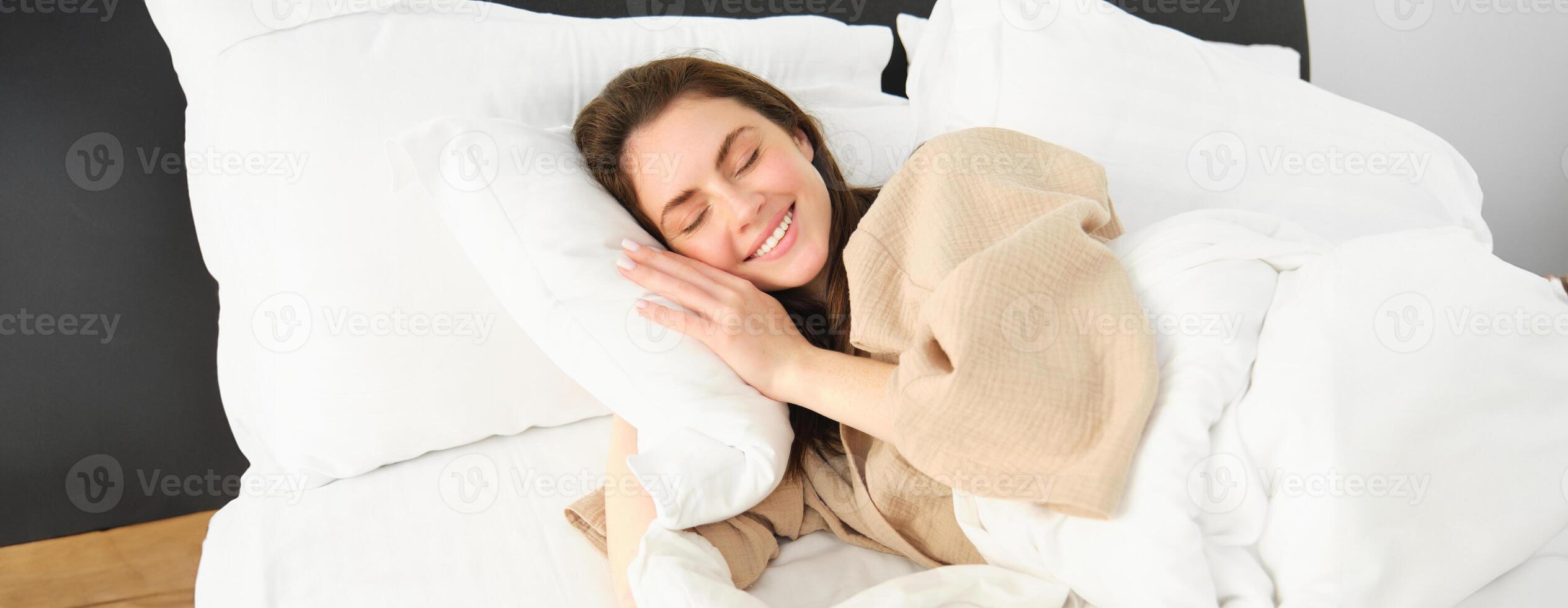 Portrait of smiling brunette woman in pyjamas, sleeping in hotel bed, relaxing with pleased face, dreaming, sleeping in bedroom photo