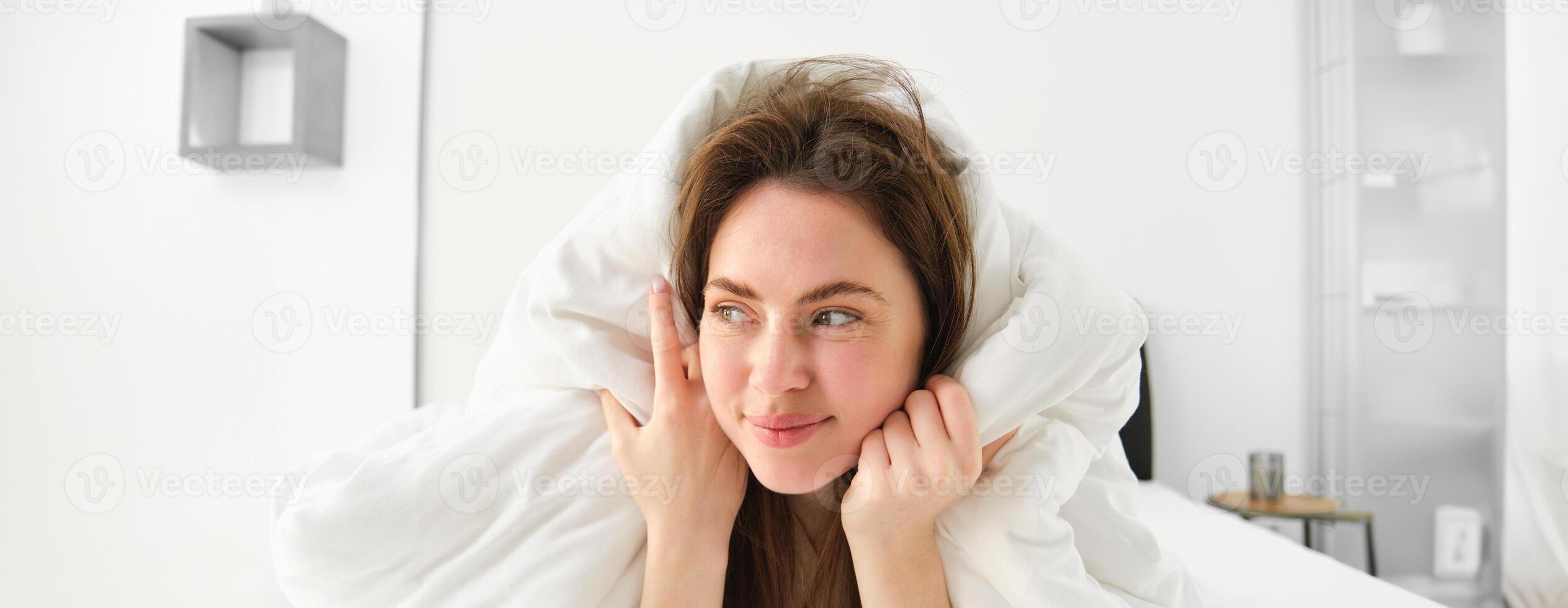 Cute girl with messy hair, lying in bed covered in white sheets duvet, smiling and laughing coquettish, spending time in her bedroom photo