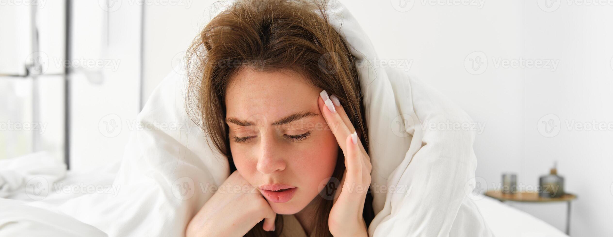 Portrait of sad woman feeling unwell, lying in bed under white blanket, touching head, has headache after hangover, has depression photo
