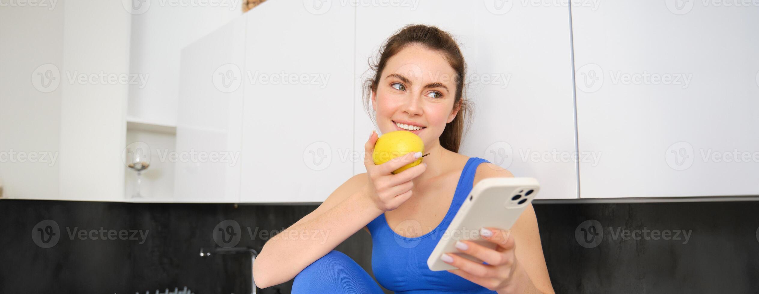 Portrait of sportswoman, girl eating and apple and looking at her social media, smartphone screen, having a snack in kitchen, wearing fitness activewear photo