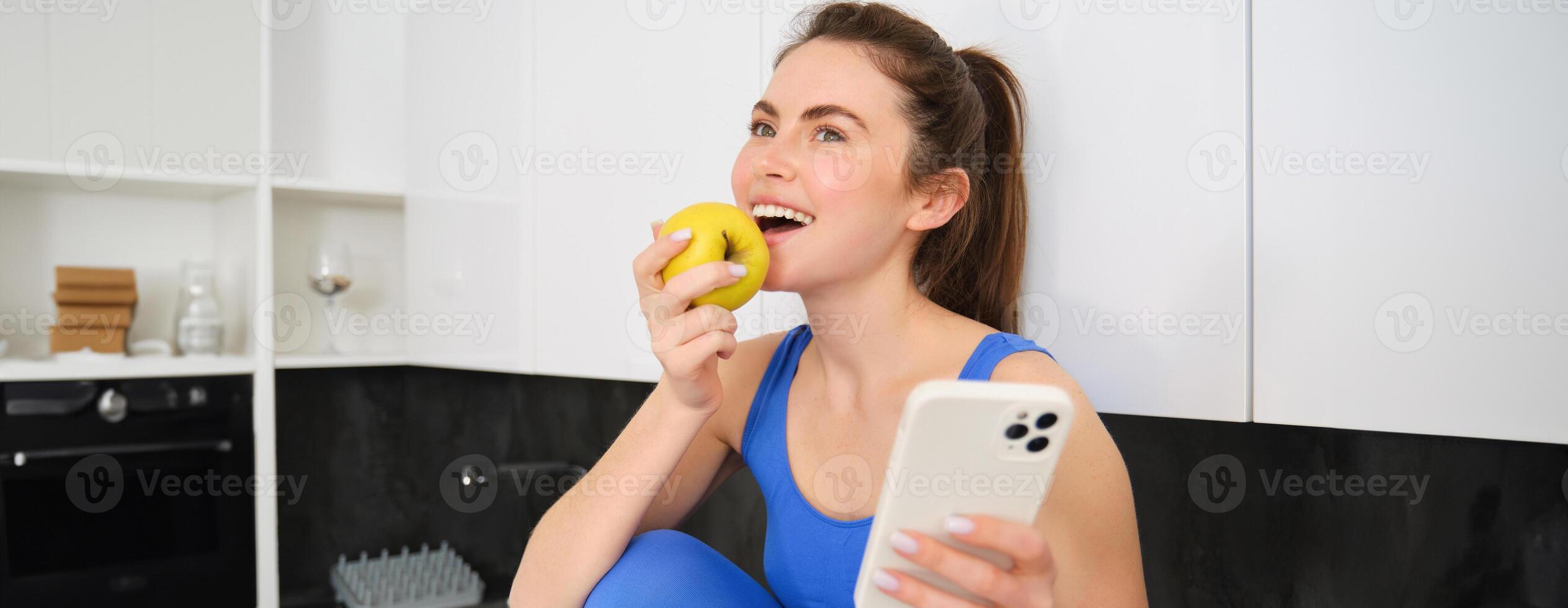 retrato de elegante, joven aptitud mujer, comiendo un manzana y utilizando móvil teléfono, participación teléfono inteligente, vistiendo ropa de deporte foto