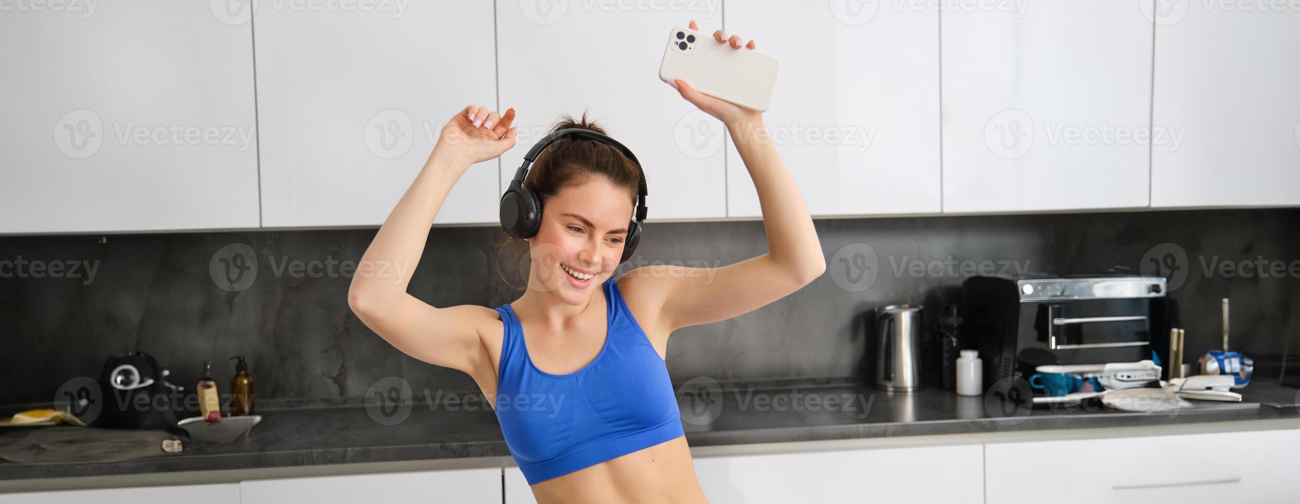 Portrait of carefree sportswoman, dancing in kitchen with smartphone, listening music in headphones, feeling energized and excited after workout photo