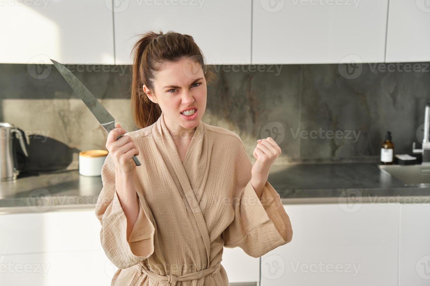 Portrait of annoyed woman with knife, angry while cooking in the kitchen, frustrated while doing house chores and preparing food for family, standing in bathrobe photo