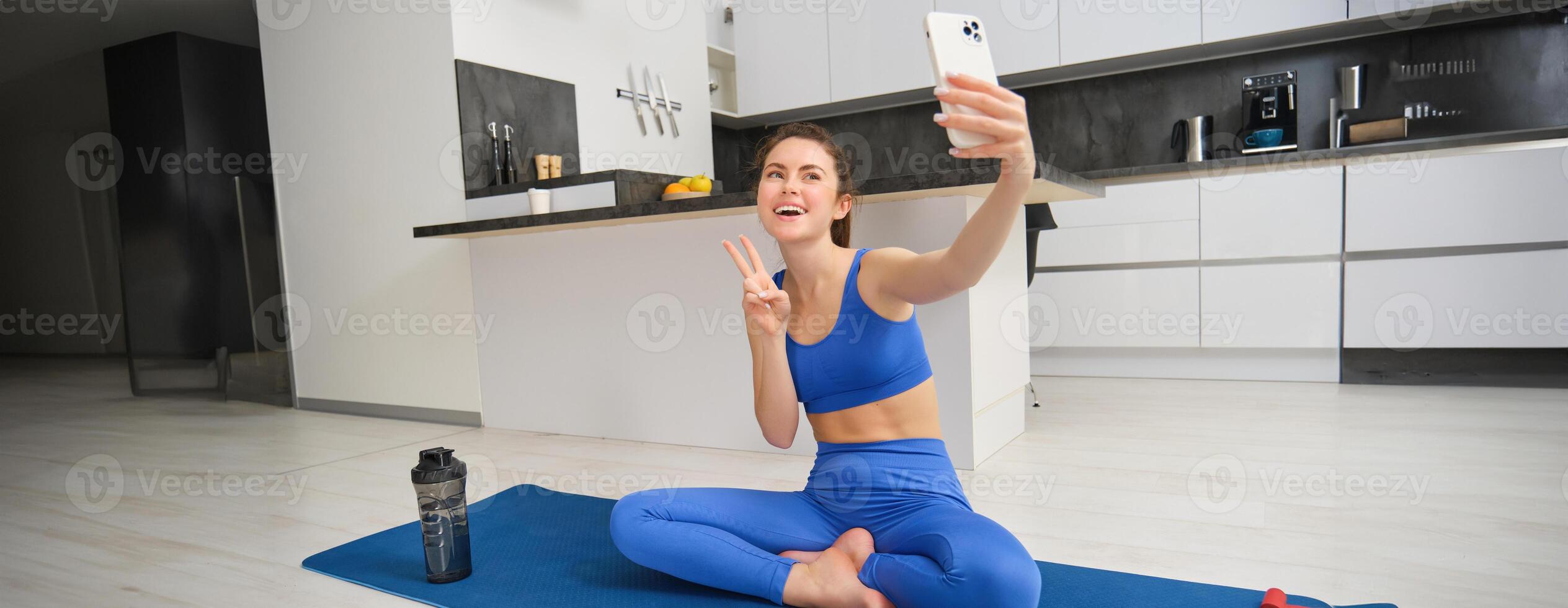 Active young woman, vlogger does sports, records her workout training from home on smartphone camera, posing for selfie inside her house, sits on rubber yoga mat in blue leggings and sportsbra photo