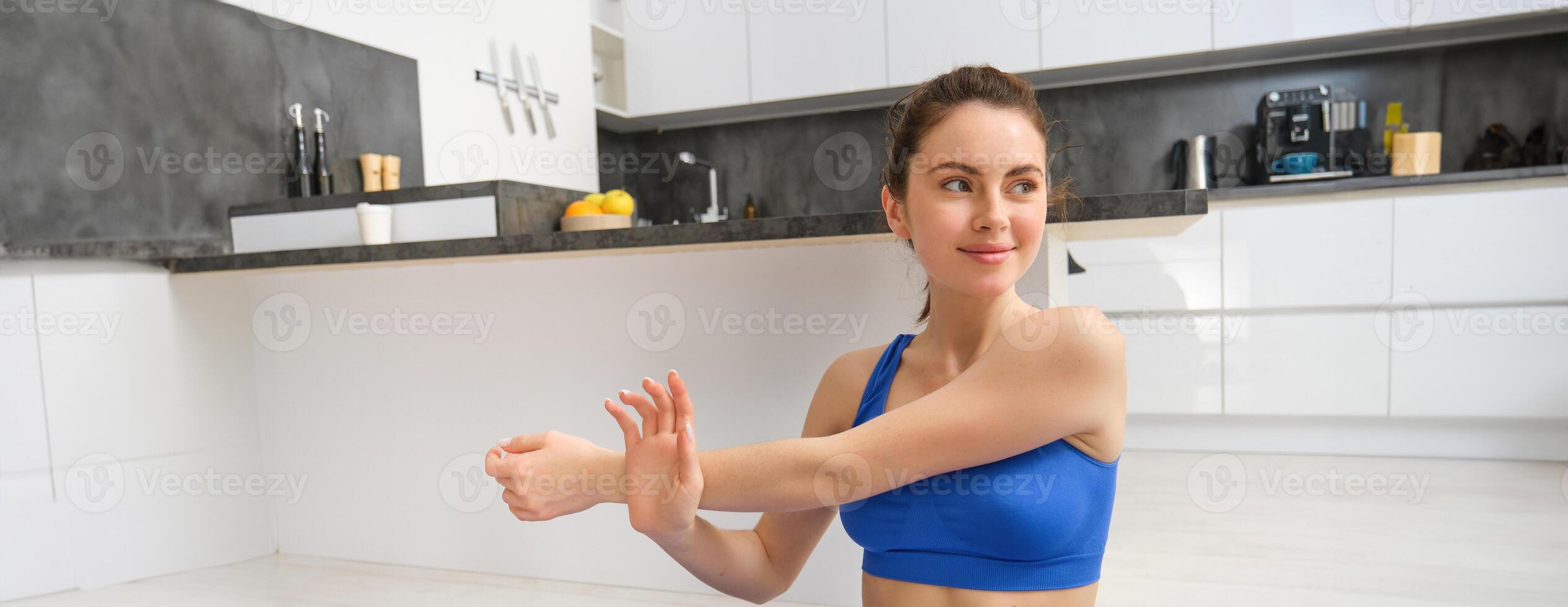 hermosa y ajuste mujer lo hace rutina de ejercicio a hogar, estiramientos su brazos, lo hace calentar antes de formación ejercicios, usa sujetador deportivo, se sienta en caucho yoga estera foto