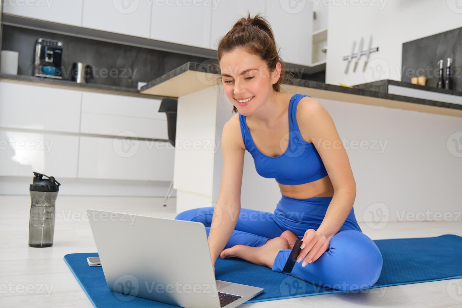 Portrait of young woman following online fitness lesson at home, sitting on yoga mat and using laptop during aerobics workout photo