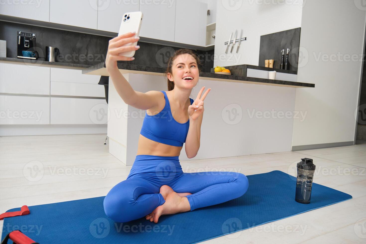 activo joven mujer, vlogger lo hace Deportes, registros su rutina de ejercicio formación desde hogar en teléfono inteligente cámara, posando para selfie dentro su casa, se sienta en caucho yoga estera en azul polainas y sujetador deportivo foto