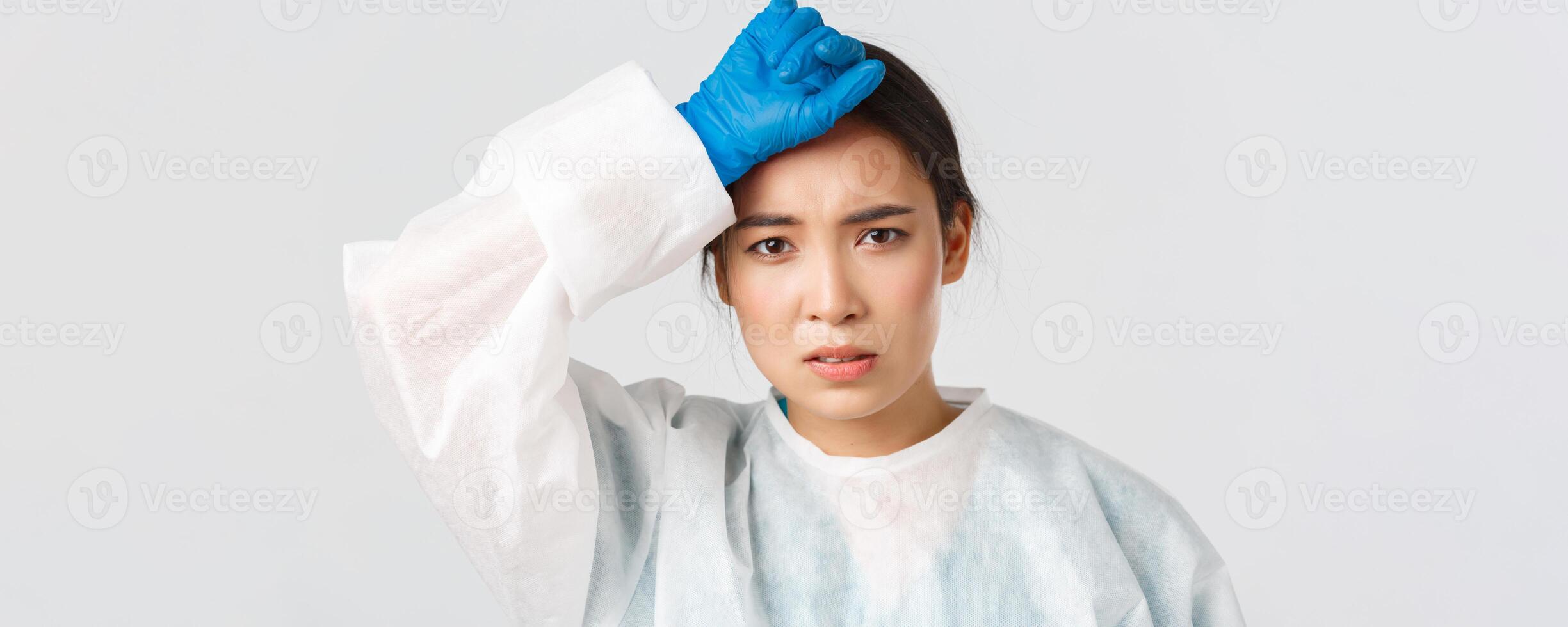 Covid-19, coronavirus disease, healthcare workers concept. Young tired asian female doctor take-off personal protective equipment and wipe sweat, looking exhausted after long shift in hospital photo