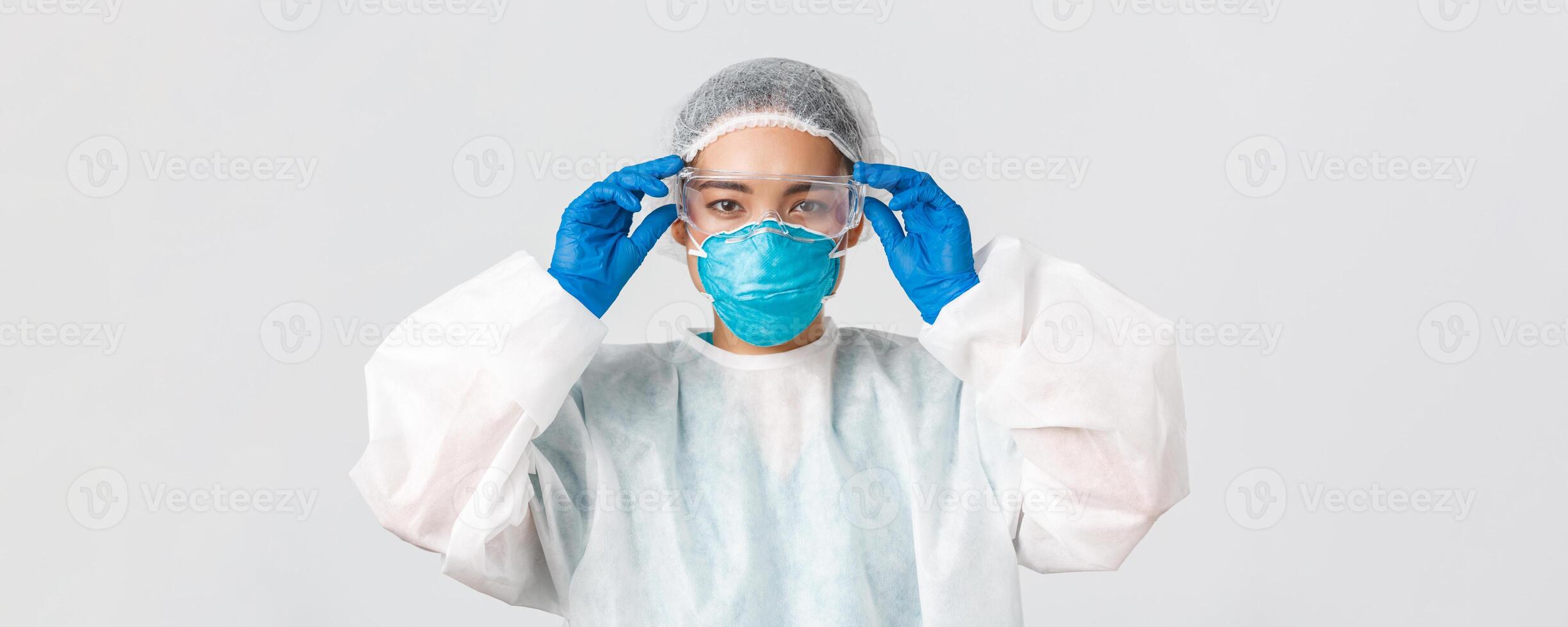 Covid-19, coronavirus disease, healthcare workers concept. Serious-looking asian female doctor in personal protective equipment, put on glass before entering lab, white background photo