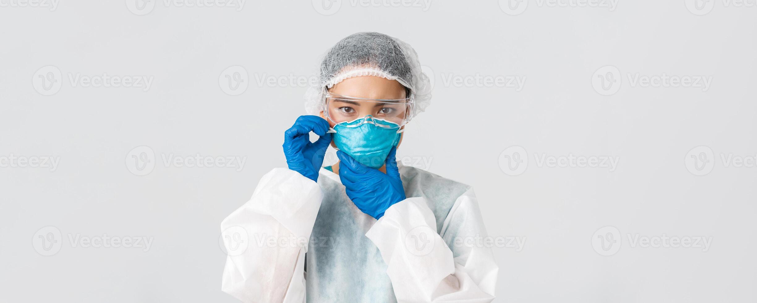 Covid-19, coronavirus disease, healthcare workers concept. Serious asian female doctor prepare enter contagious zone, examine sick patients, put on PPE and respirator, white background photo