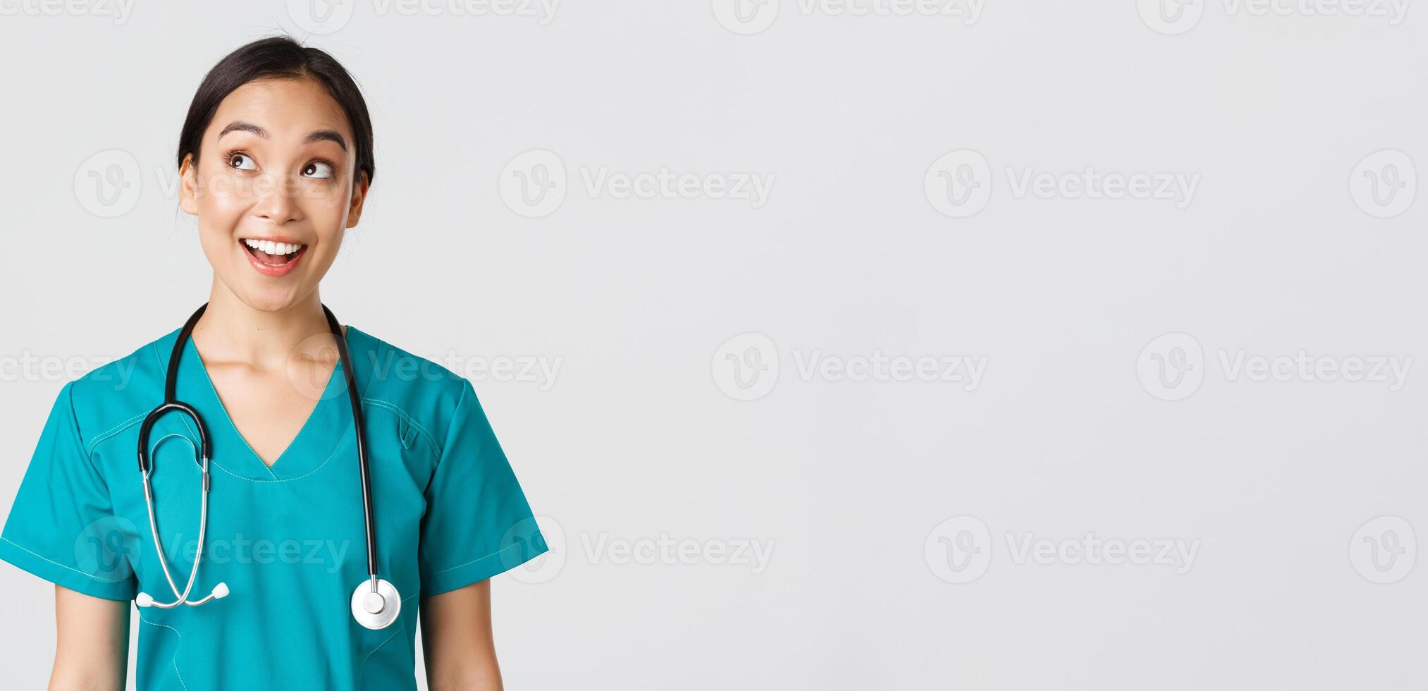Healthcare workers, preventing virus, quarantine campaign concept. Enthusiastic smiling asian doctor in scrubs, nurse looking upper left corner with amused face, standing white background photo