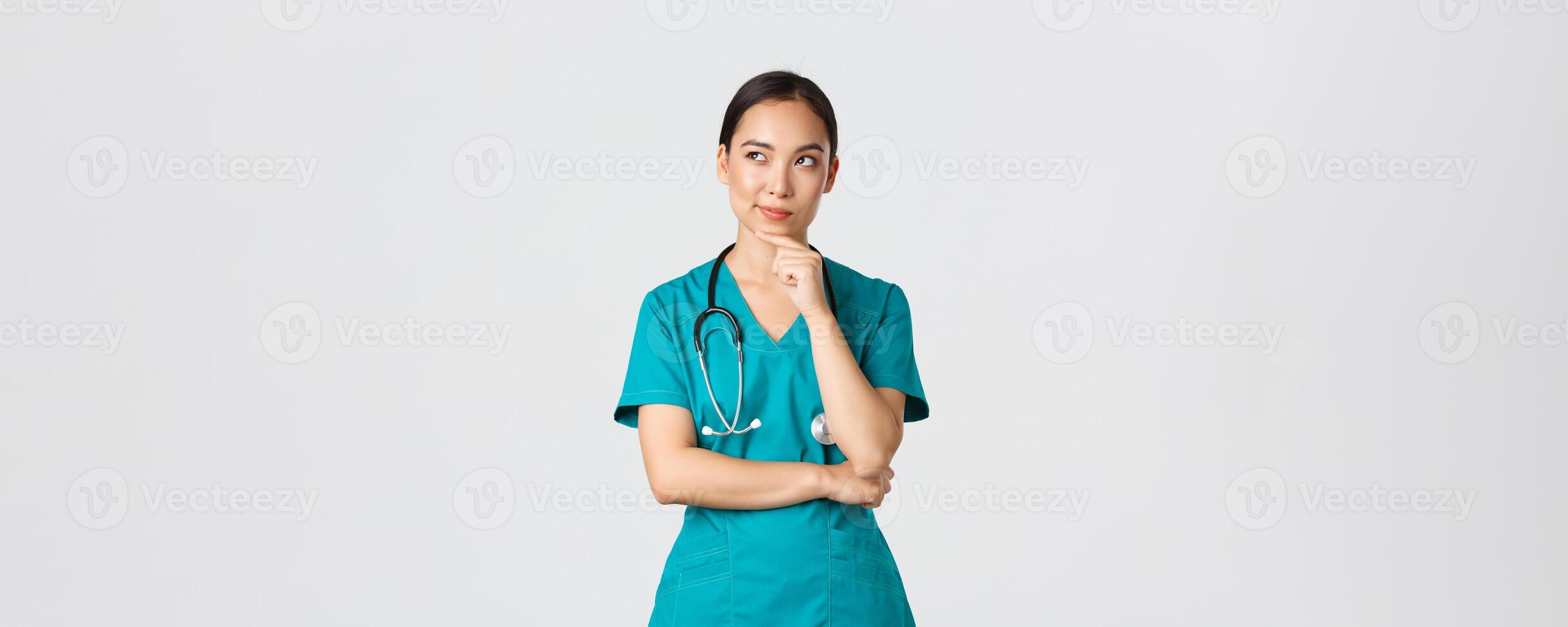 Covid-19, healthcare workers, pandemic concept. Thoughtful smart asian nurse in scrubs looking away and thinking, smiling pleased. Doctor have interesting idea, pondering over white background photo