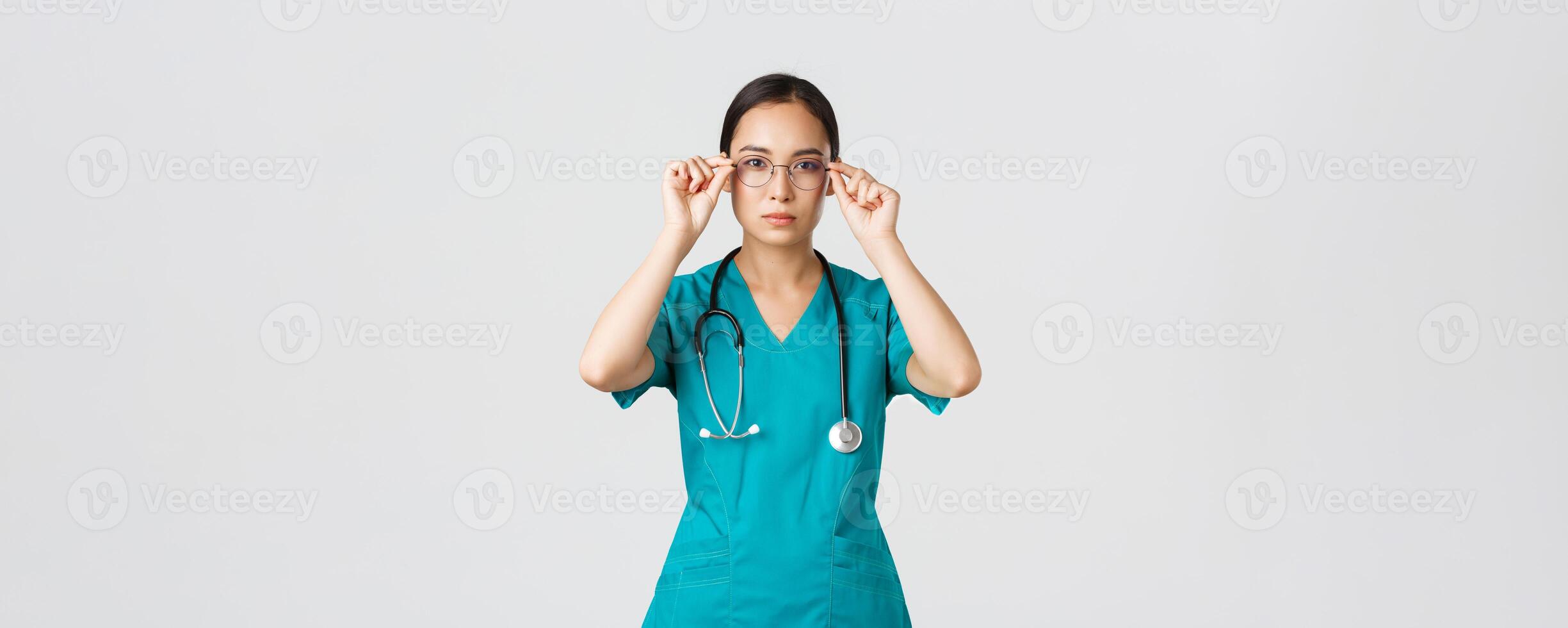 Covid-19, healthcare workers, pandemic concept. Confident serious-looking and determined asian female doctor, nurse in scrubs put on glasses, ready for shift in emergency room, white background photo