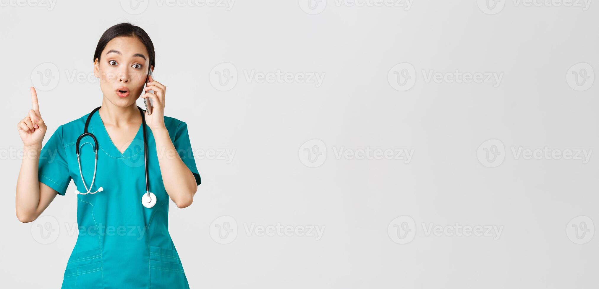 Covid-19, healthcare workers and preventing virus concept. Portrait of beautiful asian female doctor, nurse in scrubs talking on phone and raise finger as have great idea, remember, suggest plan photo