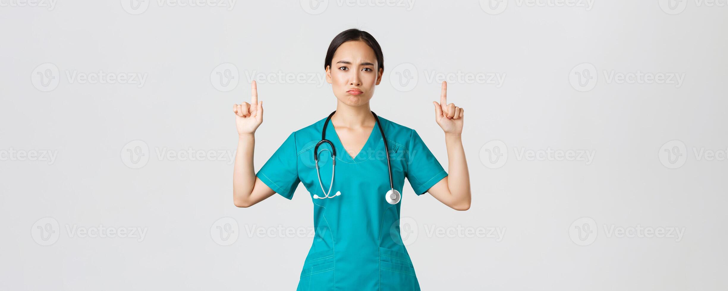 Covid-19, healthcare workers, pandemic concept. Gloomy disappointed asian female doctor, physician in scrubs pointing fingers up and grimacing displeased, standing white background photo