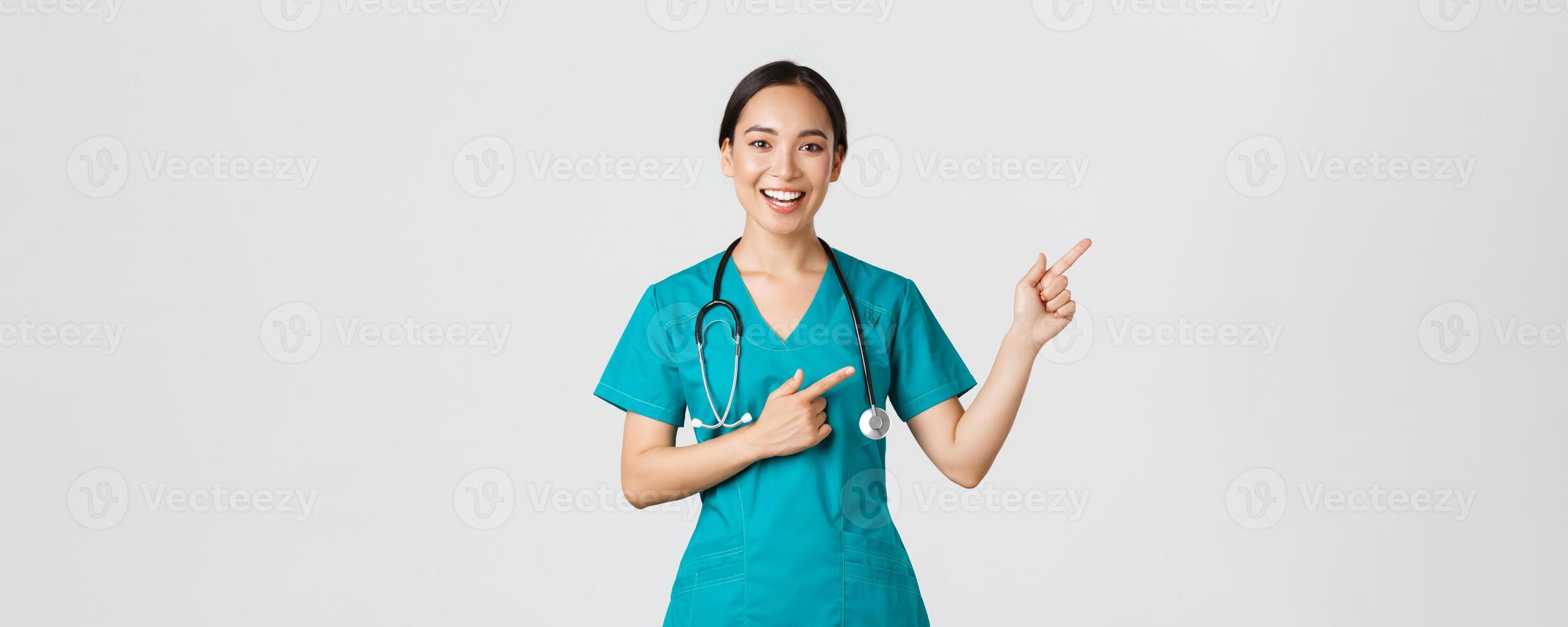 Covid-19, healthcare workers, pandemic concept. Smiling cheerful asian female doctor, therapist in scrubs pointing fingers upper right corner, inviting for checkup, recommend vaccinate photo