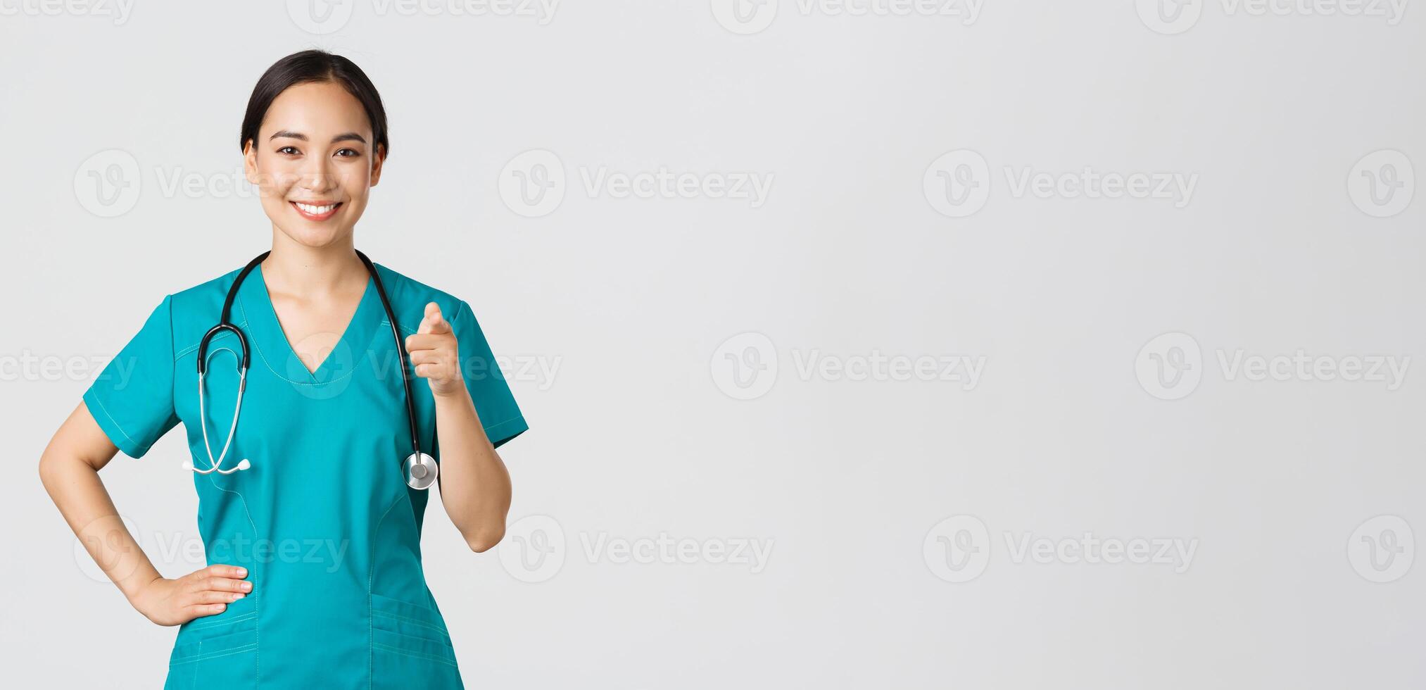 Covid-19, healthcare workers, pandemic concept. Confident smiling asian professional doctor, nurse in scrubs pointing finger at camera, inviting for check-up or vaccination, give recommendation photo