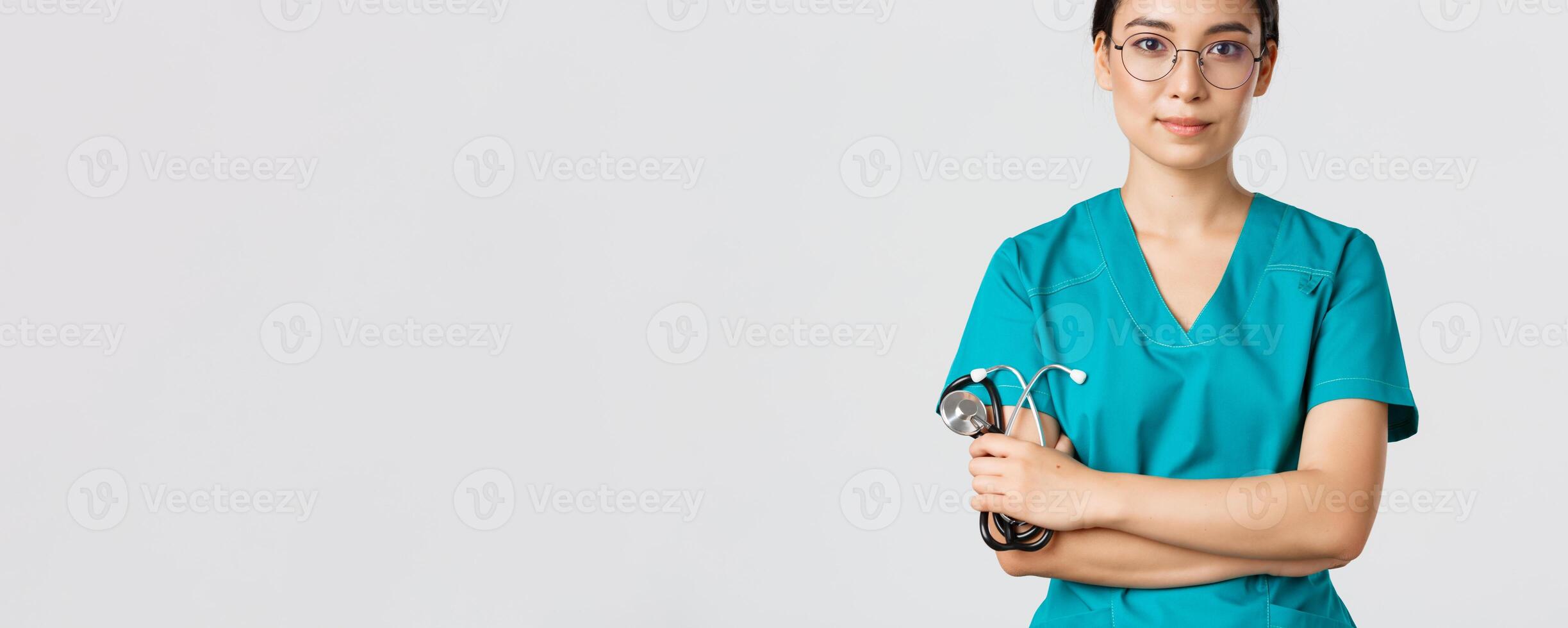 Covid-19, coronavirus disease, healthcare workers concept. Confident smiling professional asian doctor in glasses, cross arms chest, wearing scrubs and holding stethoscope, white background photo